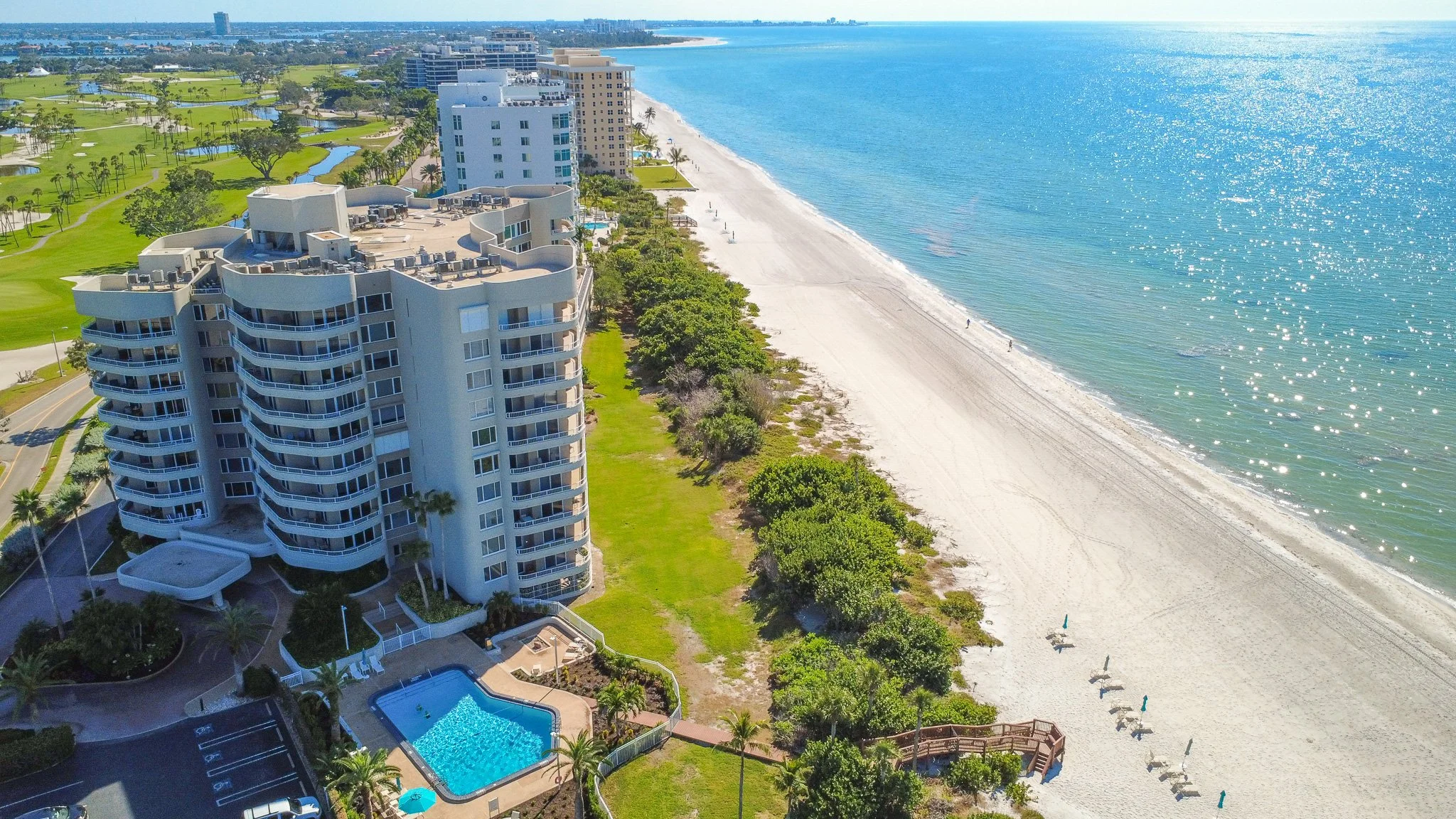 Aerial view of a beachfront hotel with a swimming pool, surrounded by greenery and tall trees, with a sandy beach and calm ocean extending into the horizon.