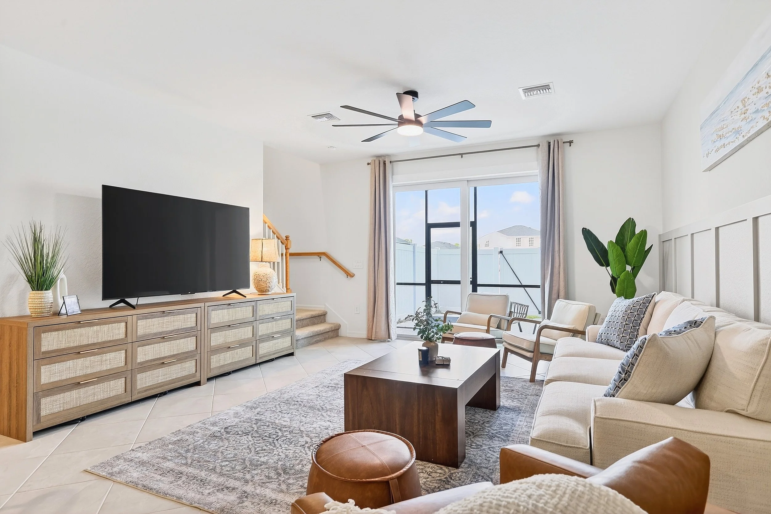 Living room with beige sofa, wooden coffee table, flat-screen TV on a wooden stand, potted plants, and a sliding glass door leading outside.