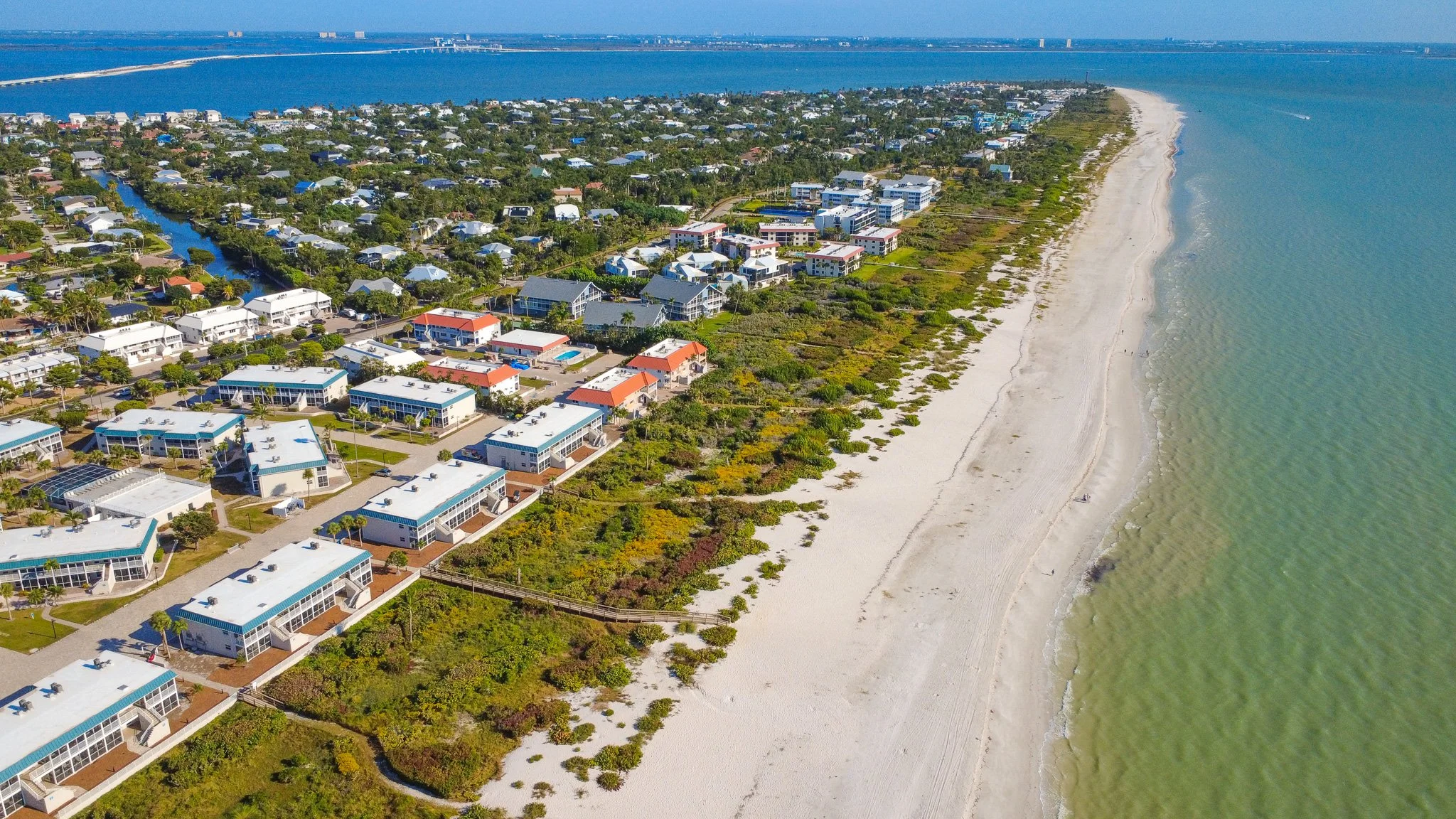 Aerial view of a coastal neighborhood with beach houses, residential homes, and a sandy shoreline next to the ocean, with a distant bridge on the water.