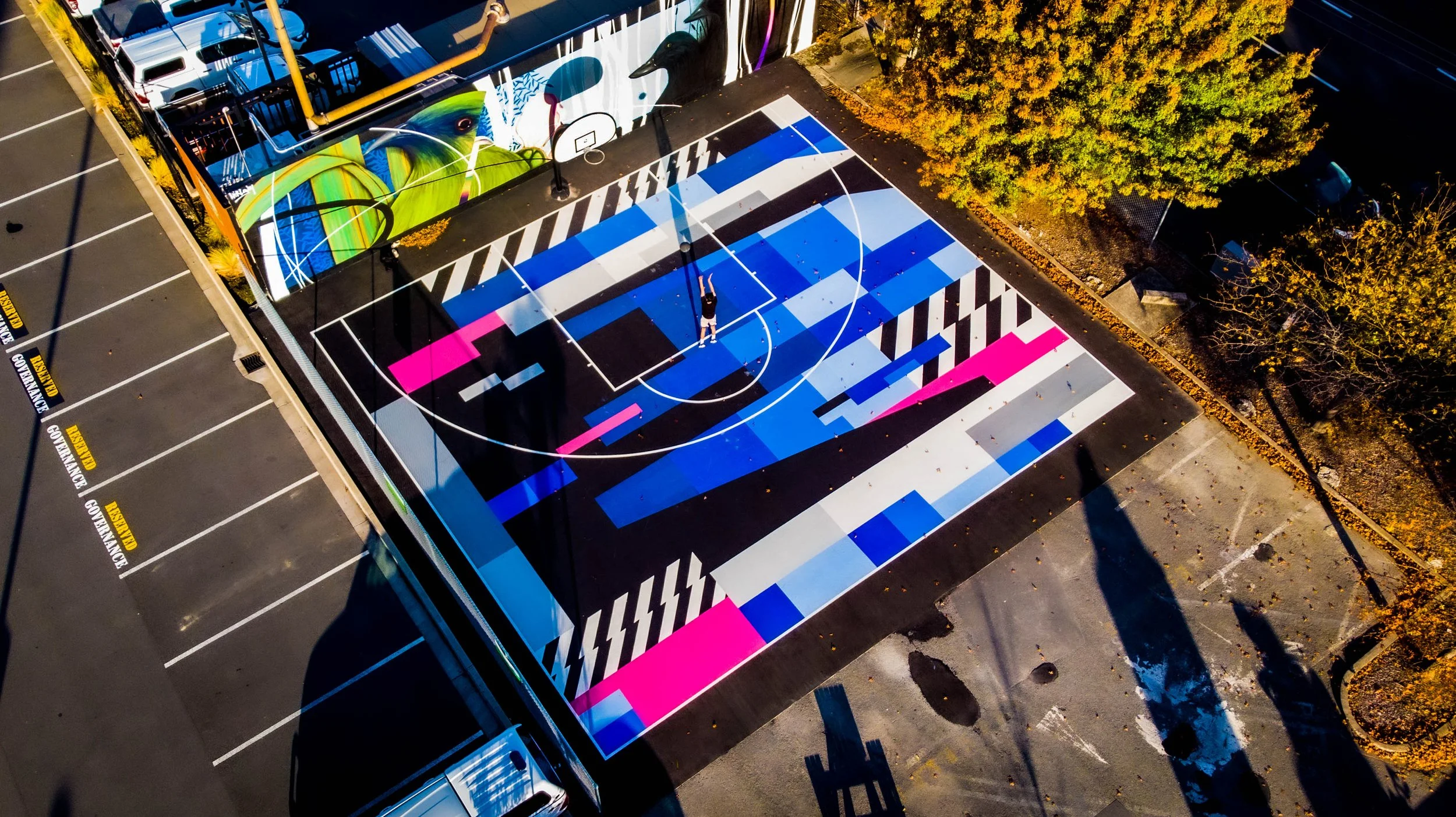 An aerial view of a colorful basketball court with geometric patterns, with a person playing at the center, surrounded by parking spaces and trees.