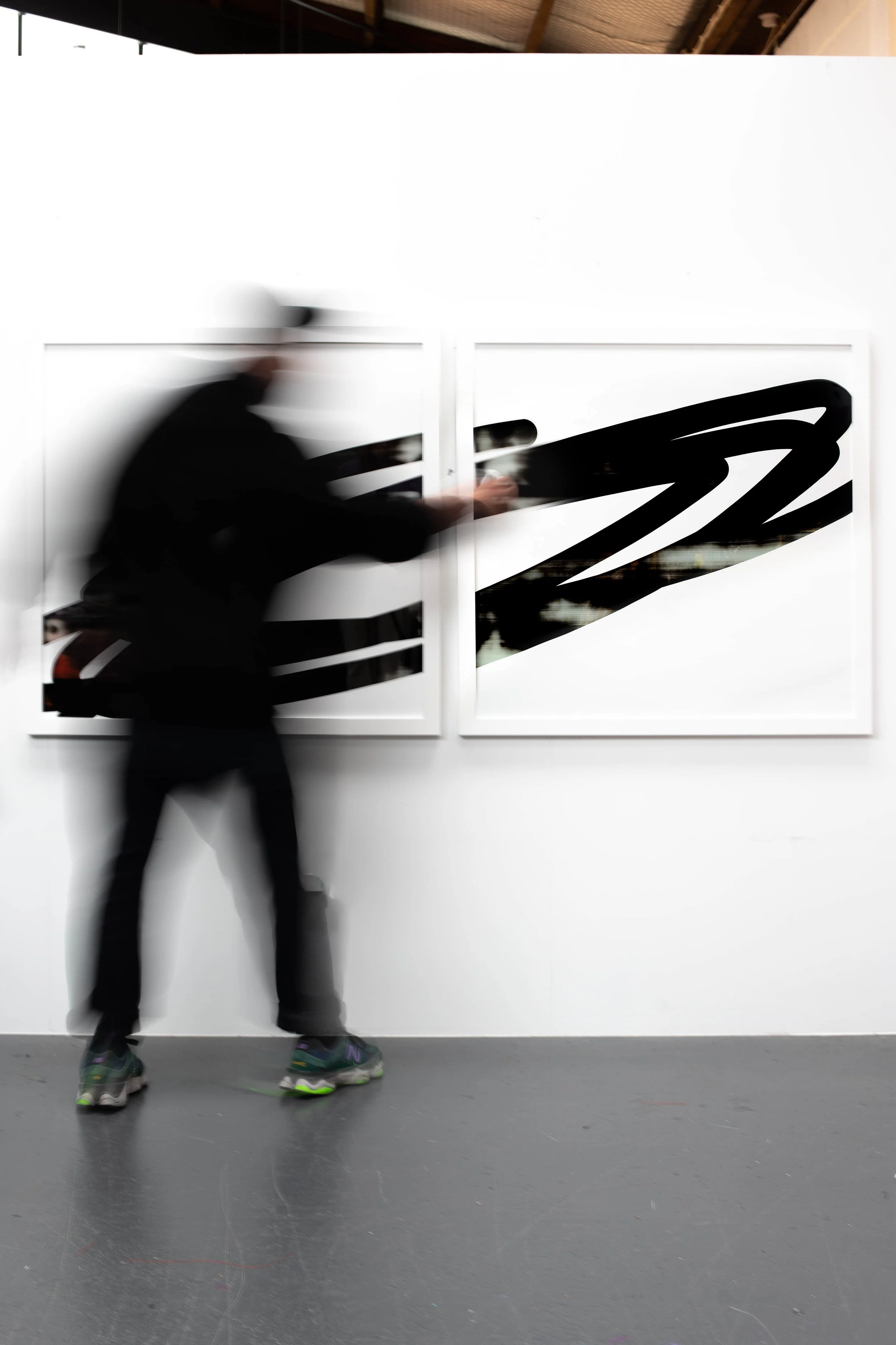 A person wearing dark clothing and sneakers is moving in front of two abstract black and white paintings on a white gallery wall, creating a motion blur effect.