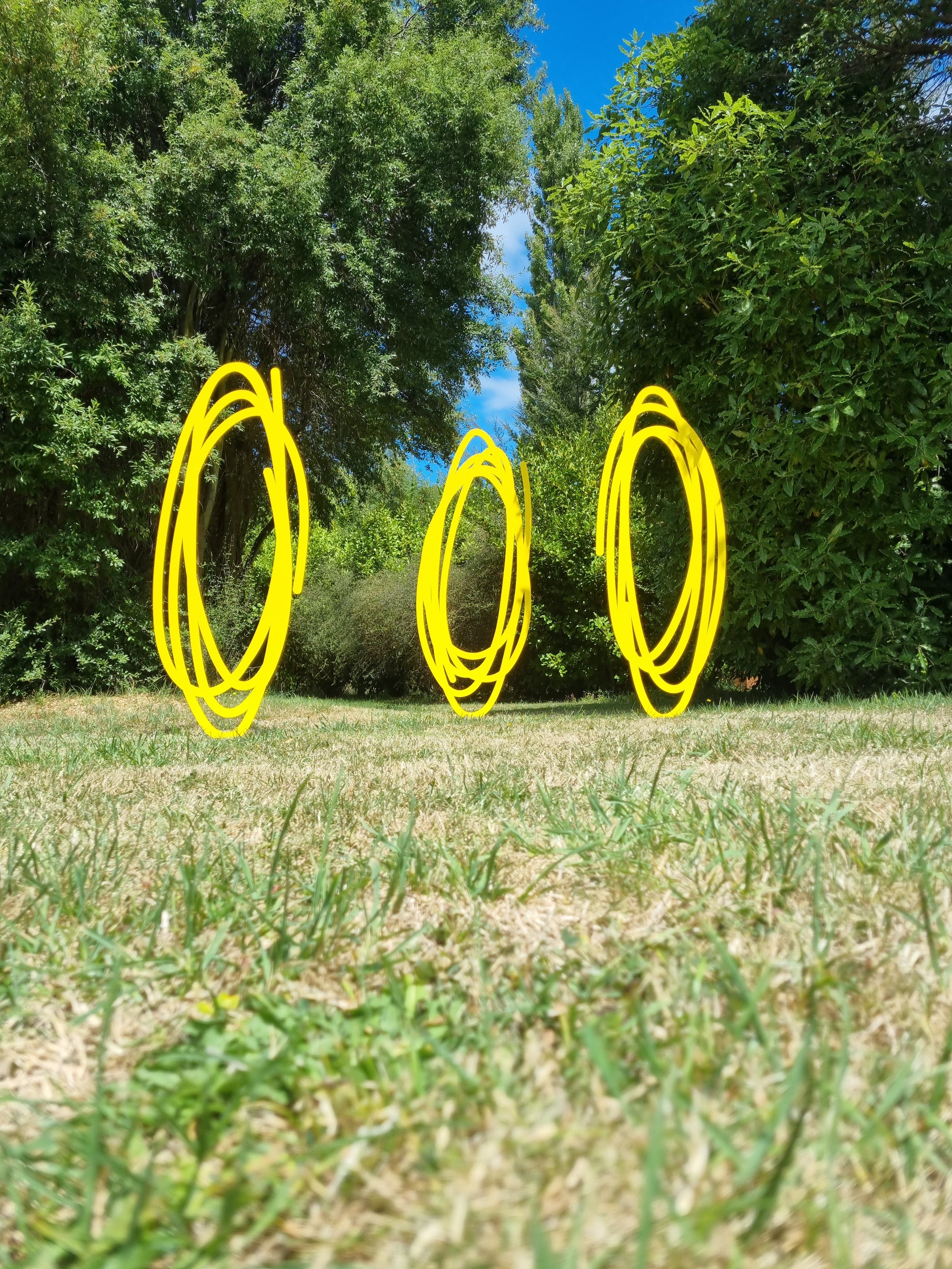 Christchurch, New Zealand artist, Nathan Ingram's three yellow, hand-drawn circle sculptures floating above the grass, surrounded by trees and blue sky.