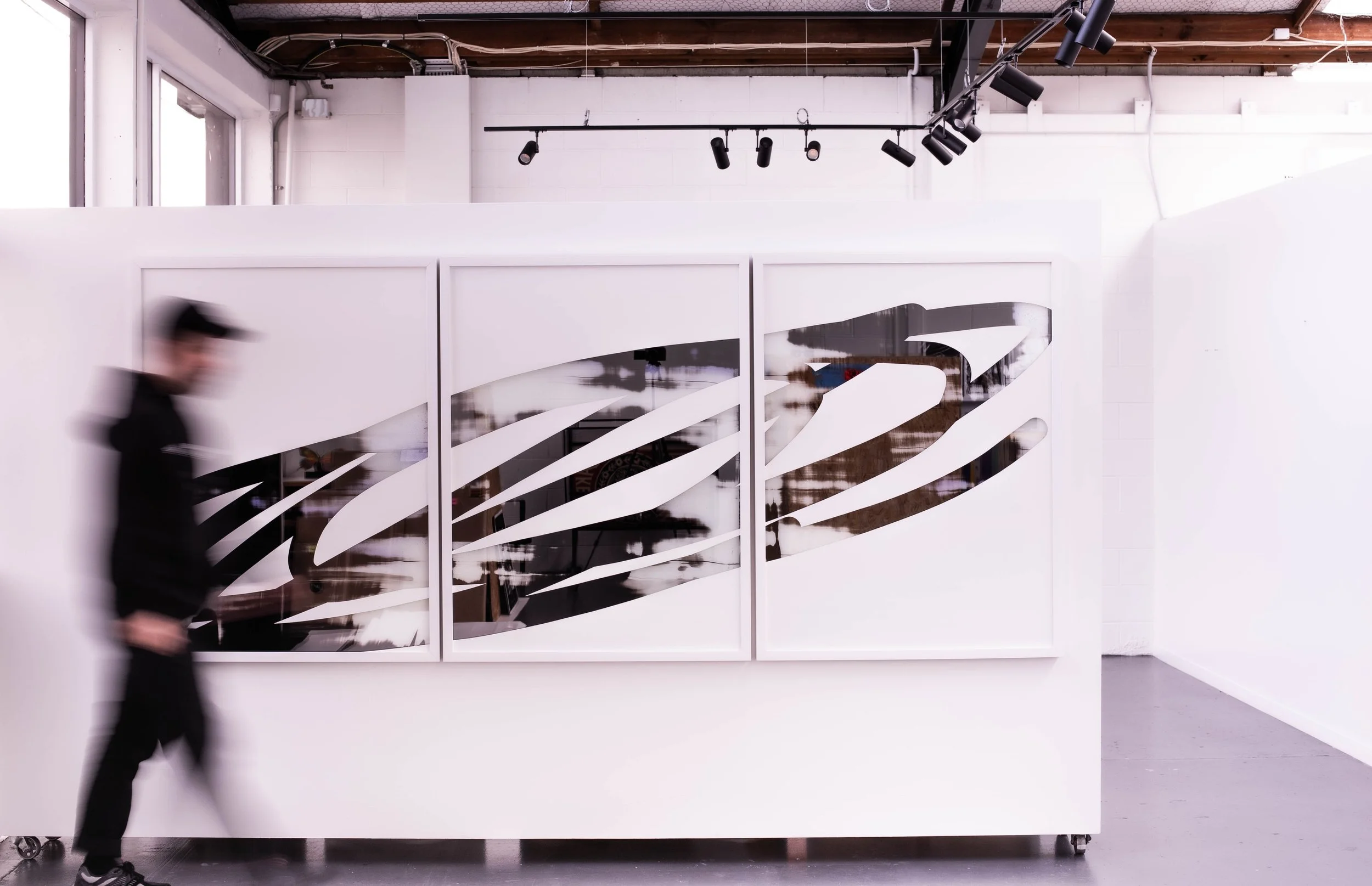 An art gallery or studio with white walls and black track lighting. Christchurch, New Zealand artist, Nathan Ingram dressed in black is walking past a white partition with three framed pieces of abstract black and white artwork.