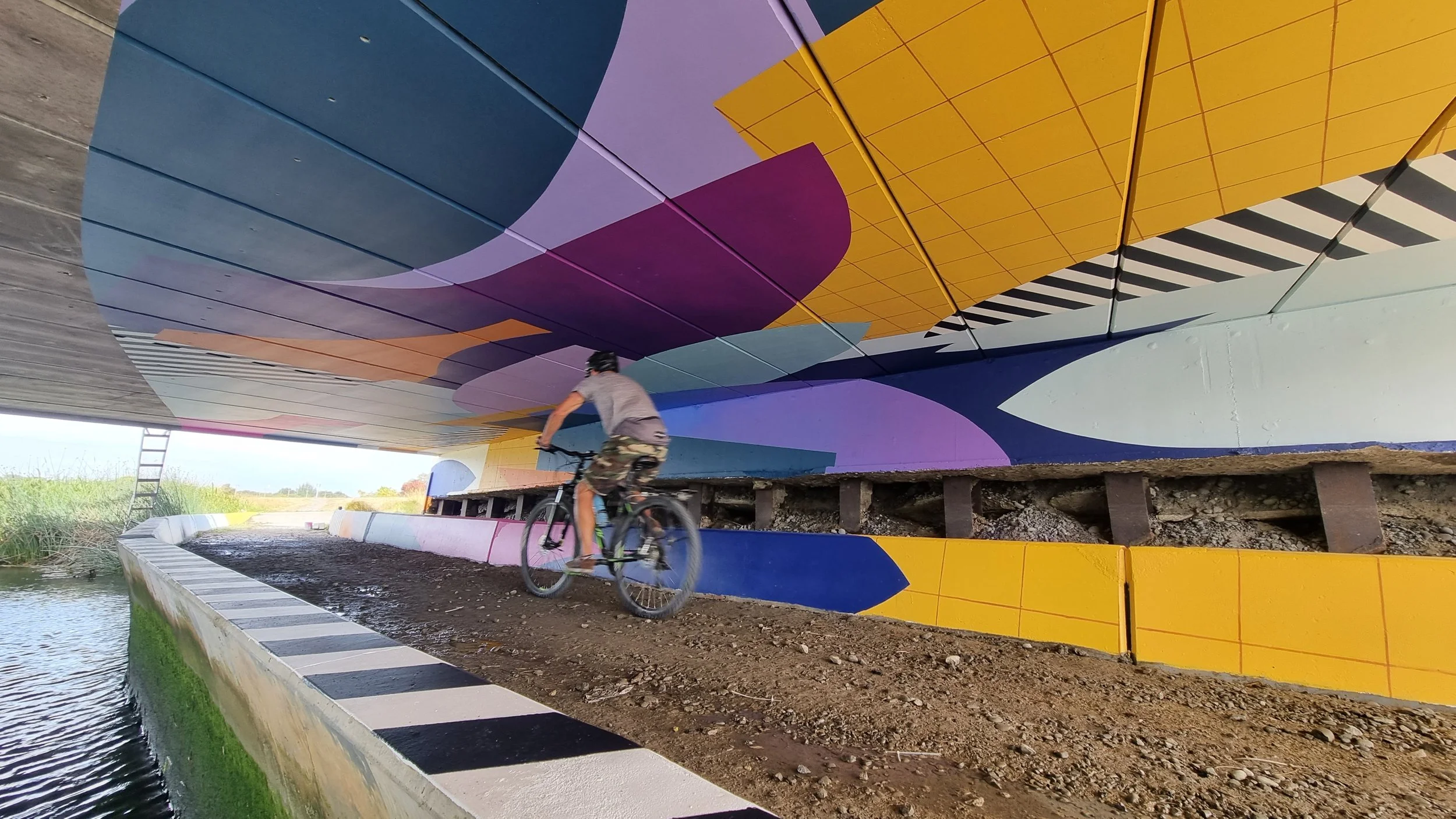 River Corridor Mural by Nathan Ingram — large-scale geometric artwork enlivening public space in Christchurch, New Zealand. A person riding a bicycle under a bridge with colorful abstract mural art on the underside.