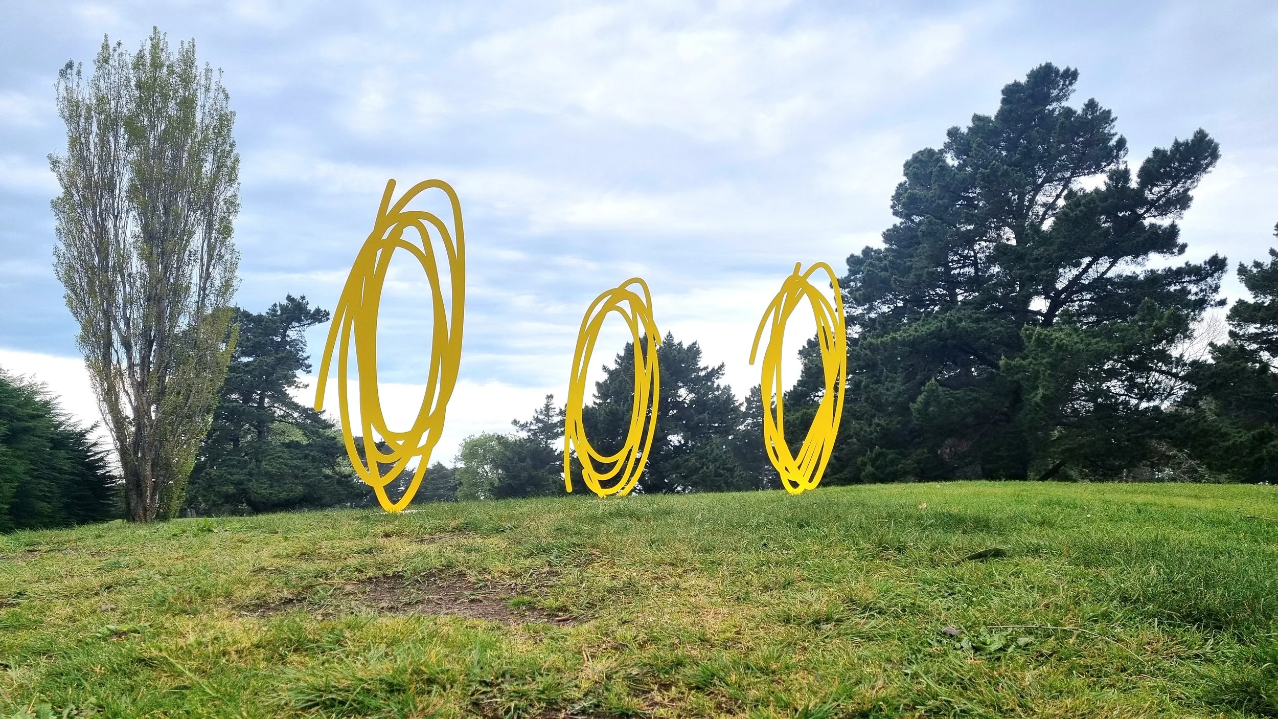 Christchurch, New Zealand artist, Nathan Ingram's three yellow, hand-drawn circle sculptures floating above the grass, surrounded by trees and blue sky.