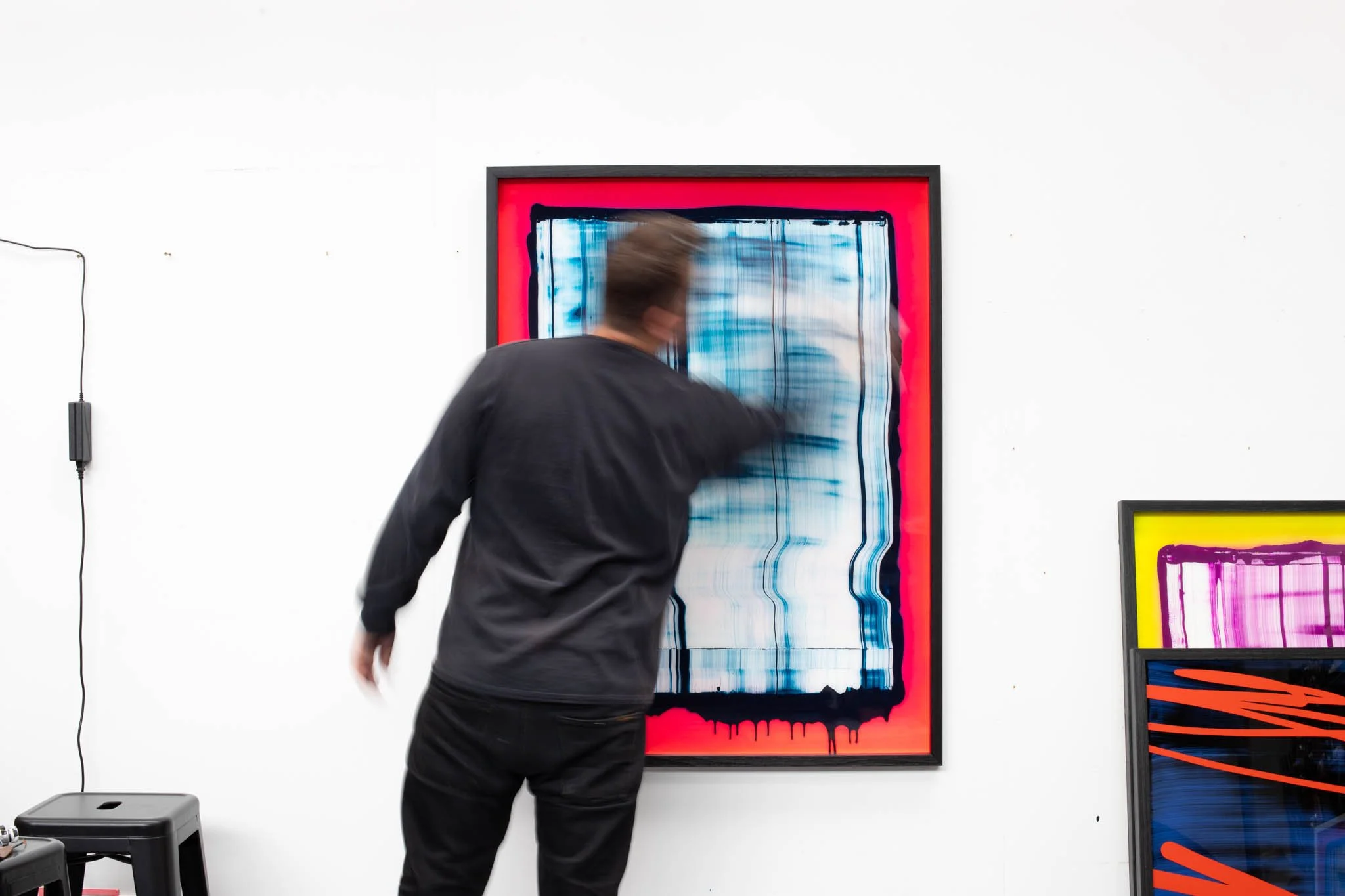 Christchurch, New Zealand artist, Nathan Ingram moving past a colorful abstract painting on a white gallery wall, with other framed artworks leaning against the wall.