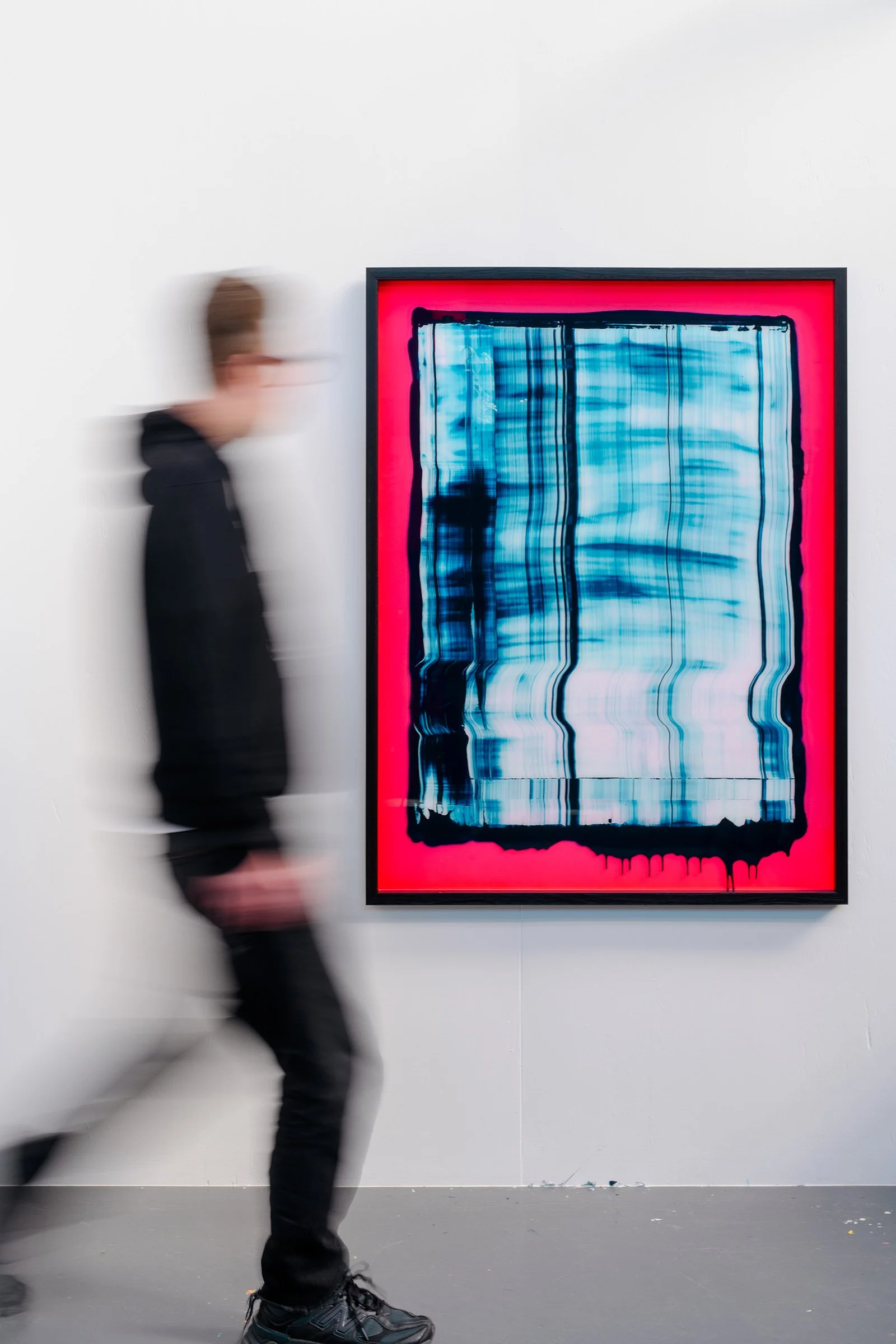 A person walking past a framed abstract painting with blue, black, and white streaks, and a red background in an art gallery.