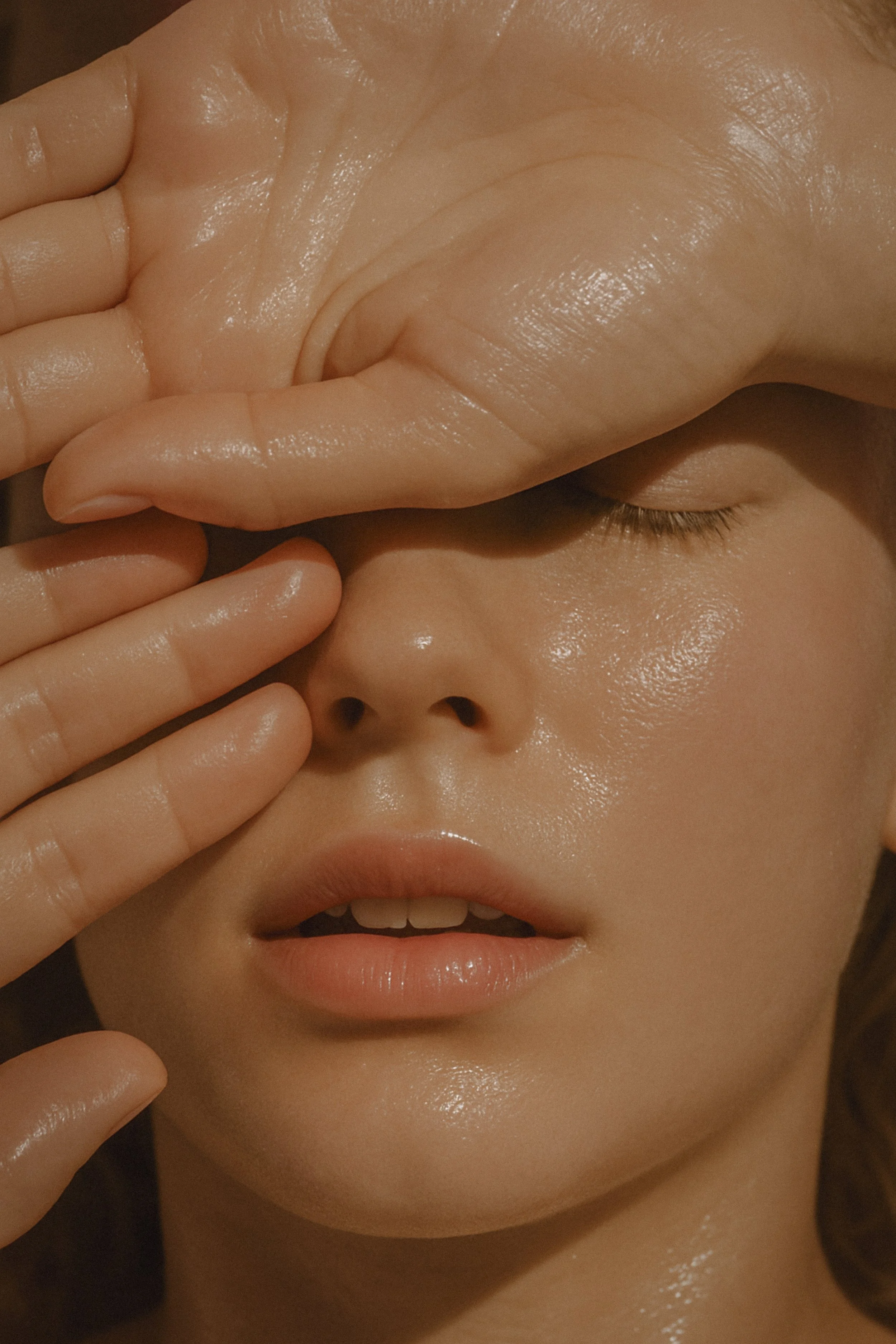 A person with closed eyes, slightly parted lips, and shiny skin, covering part of their face with their hand.