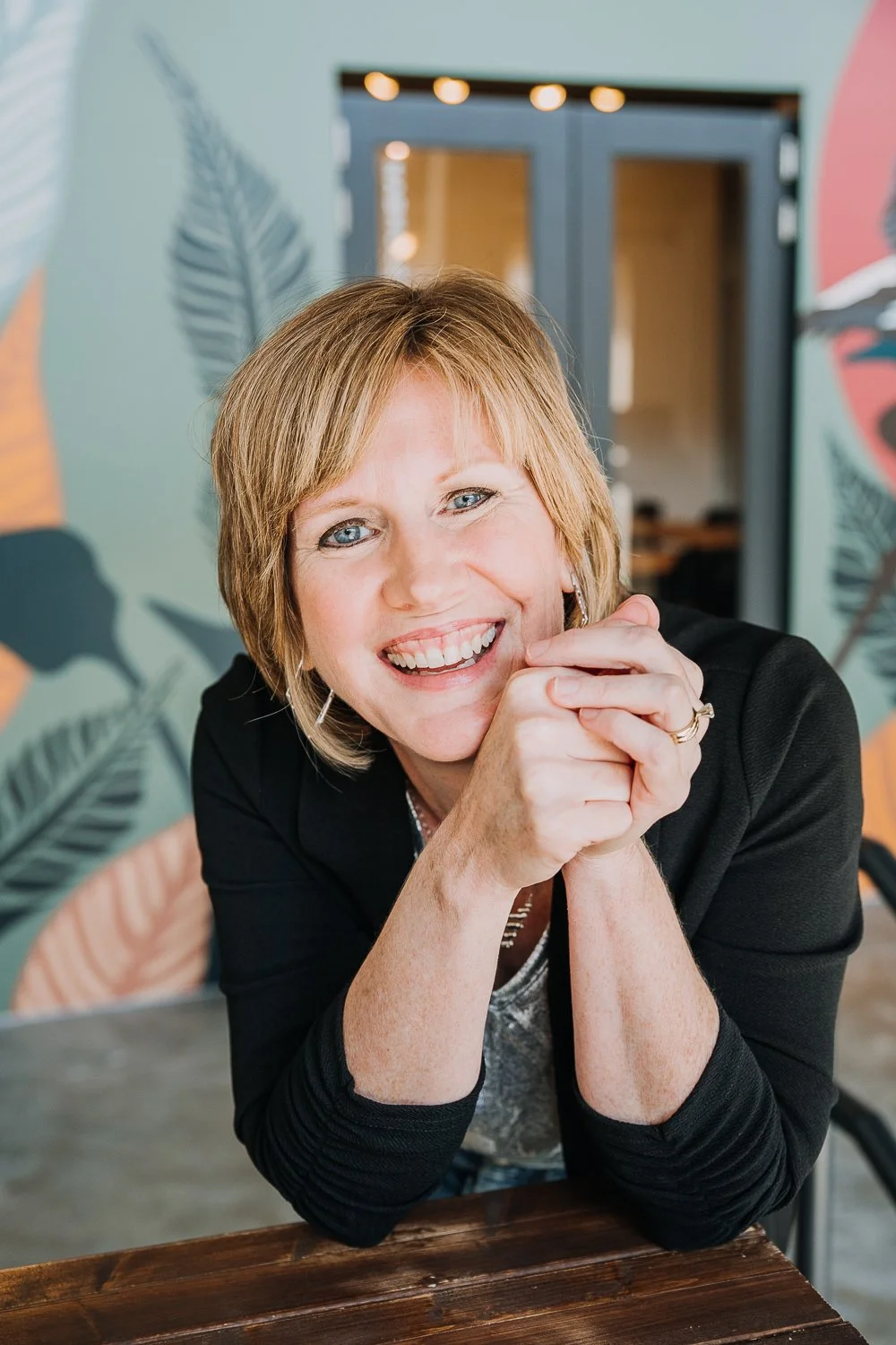 Smiling middle-aged woman with short blonde hair, wearing a black blazer and silver jewelry, sitting at a wooden table in a colorful, modern space with plant-themed wall decor.
