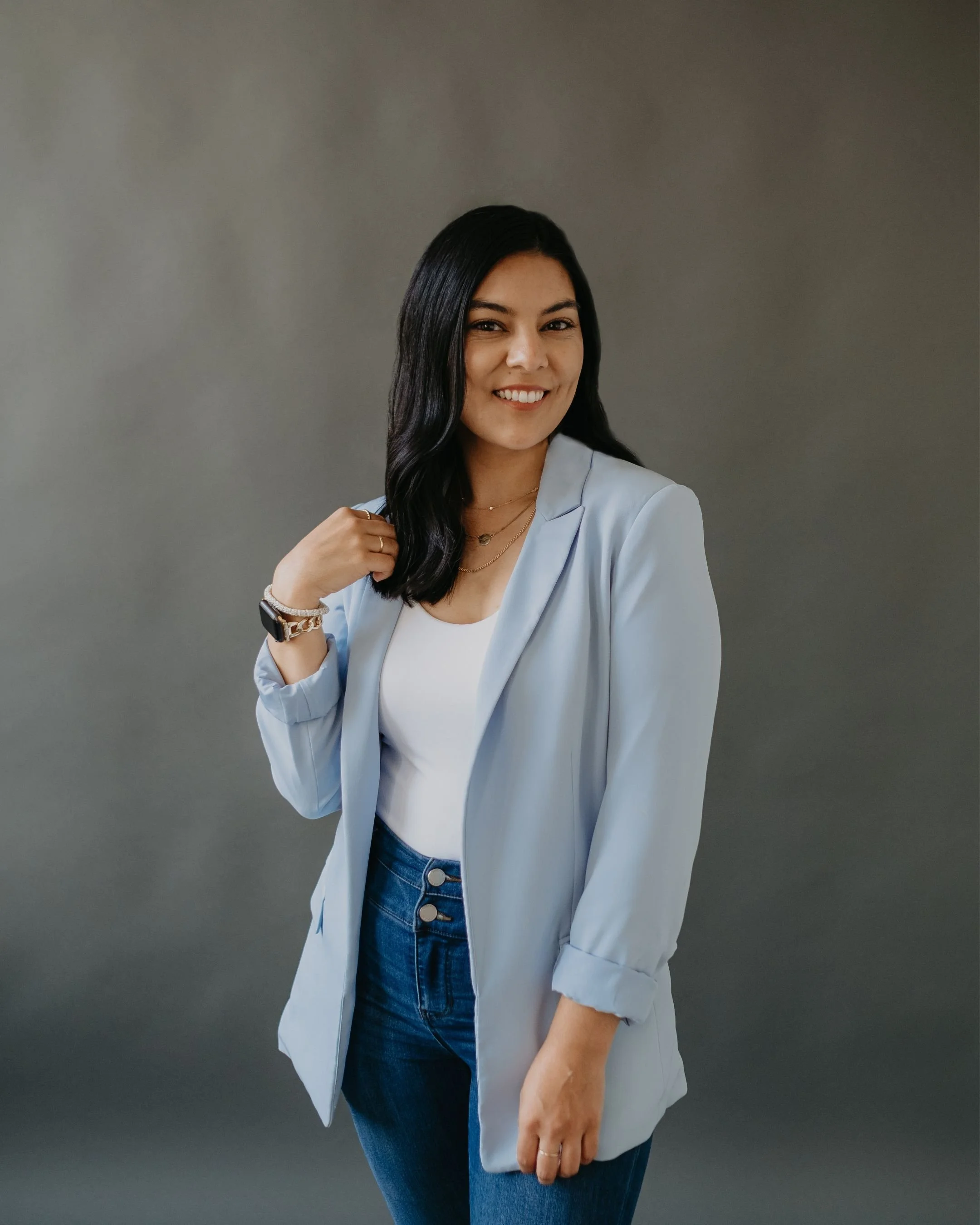 A woman with black wavy hair, smiling, wearing a light blue blazer, white top, and dark blue jeans, standing against a gray background.