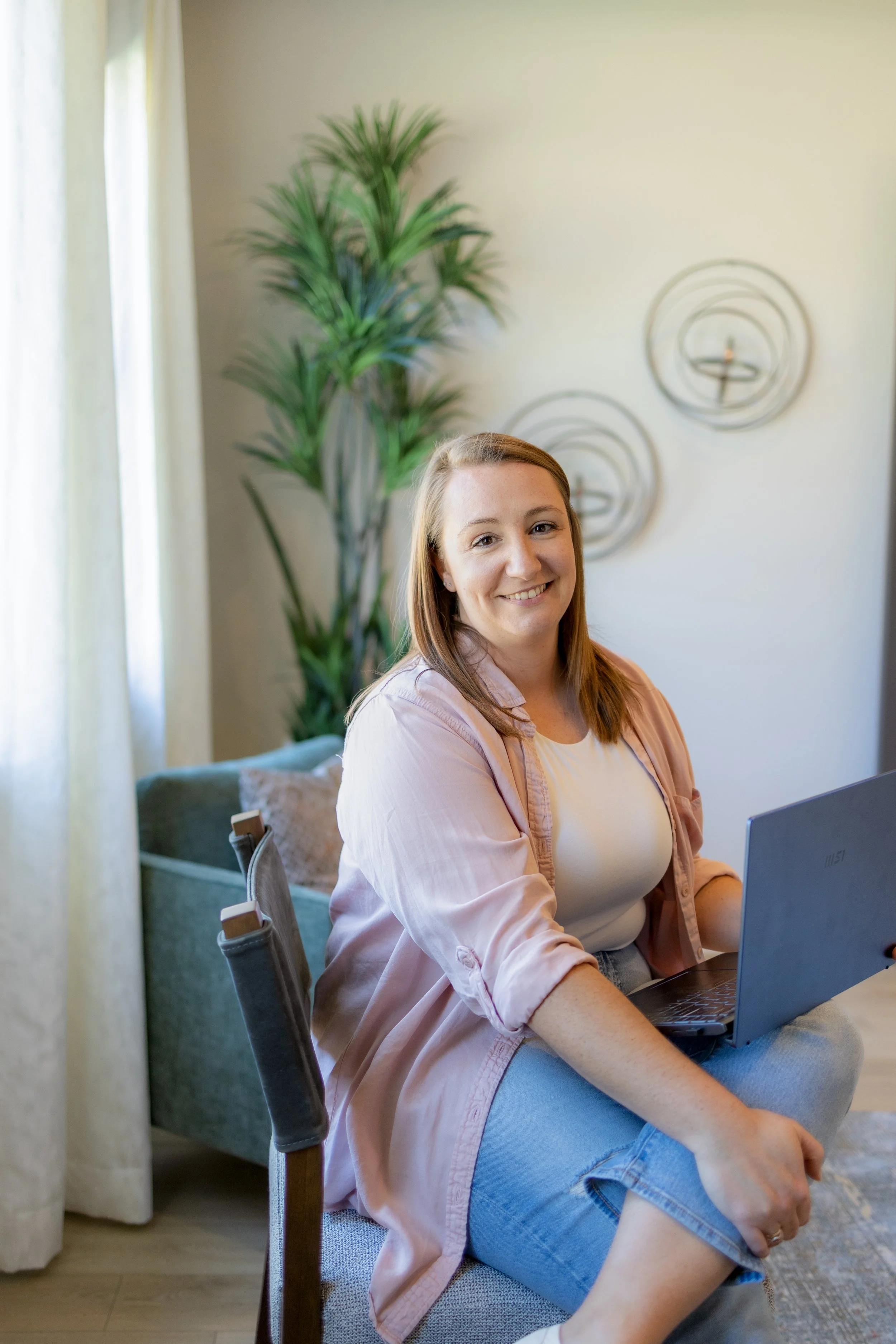 A woman sitting on a chair with a laptop on her lap, smiling at the camera, in a cozy living room with sunlight coming through curtains and a large plant in the background.