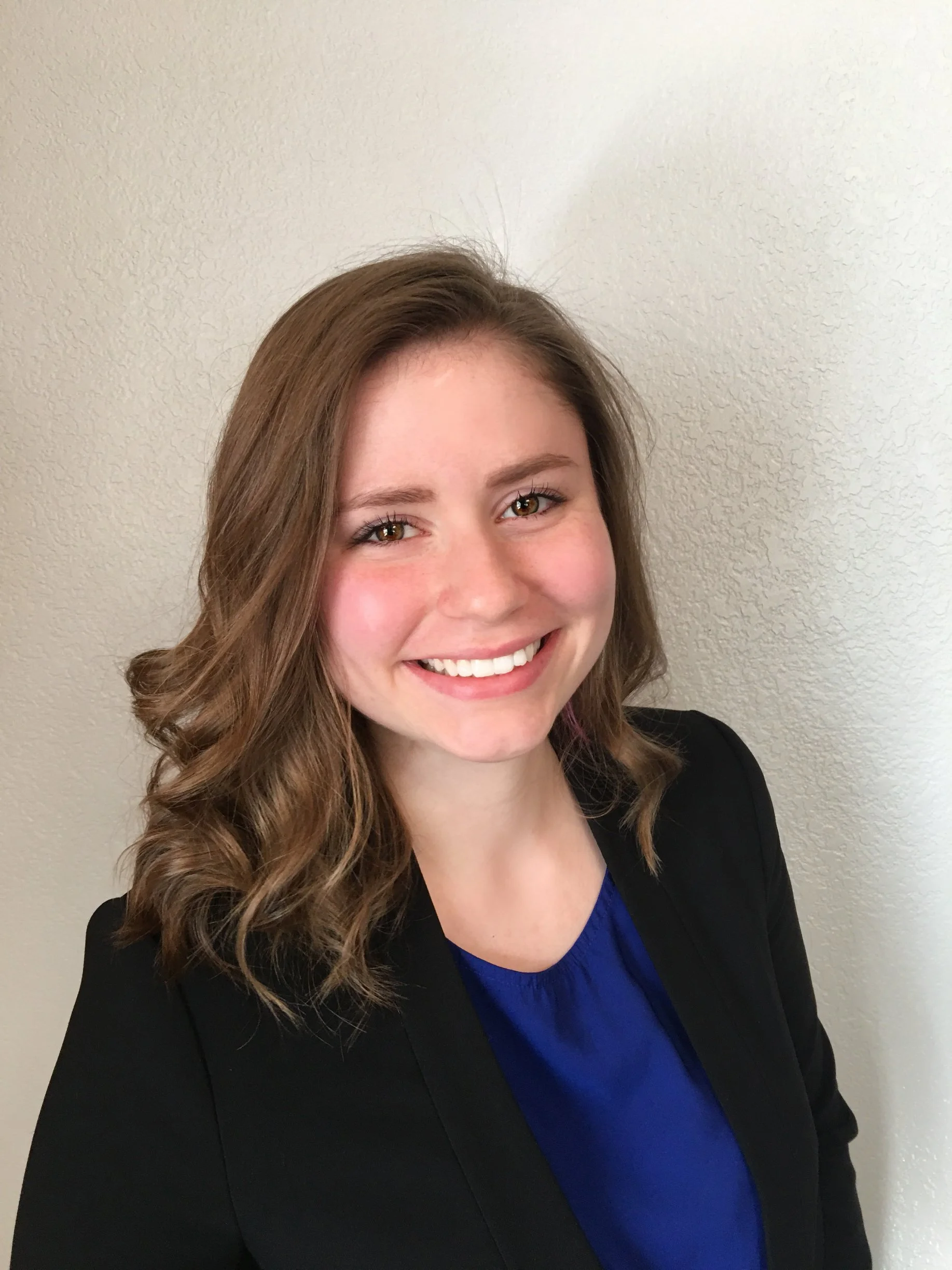 Portrait of a smiling woman with wavy brown hair wearing a black blazer and blue top against a neutral wall.