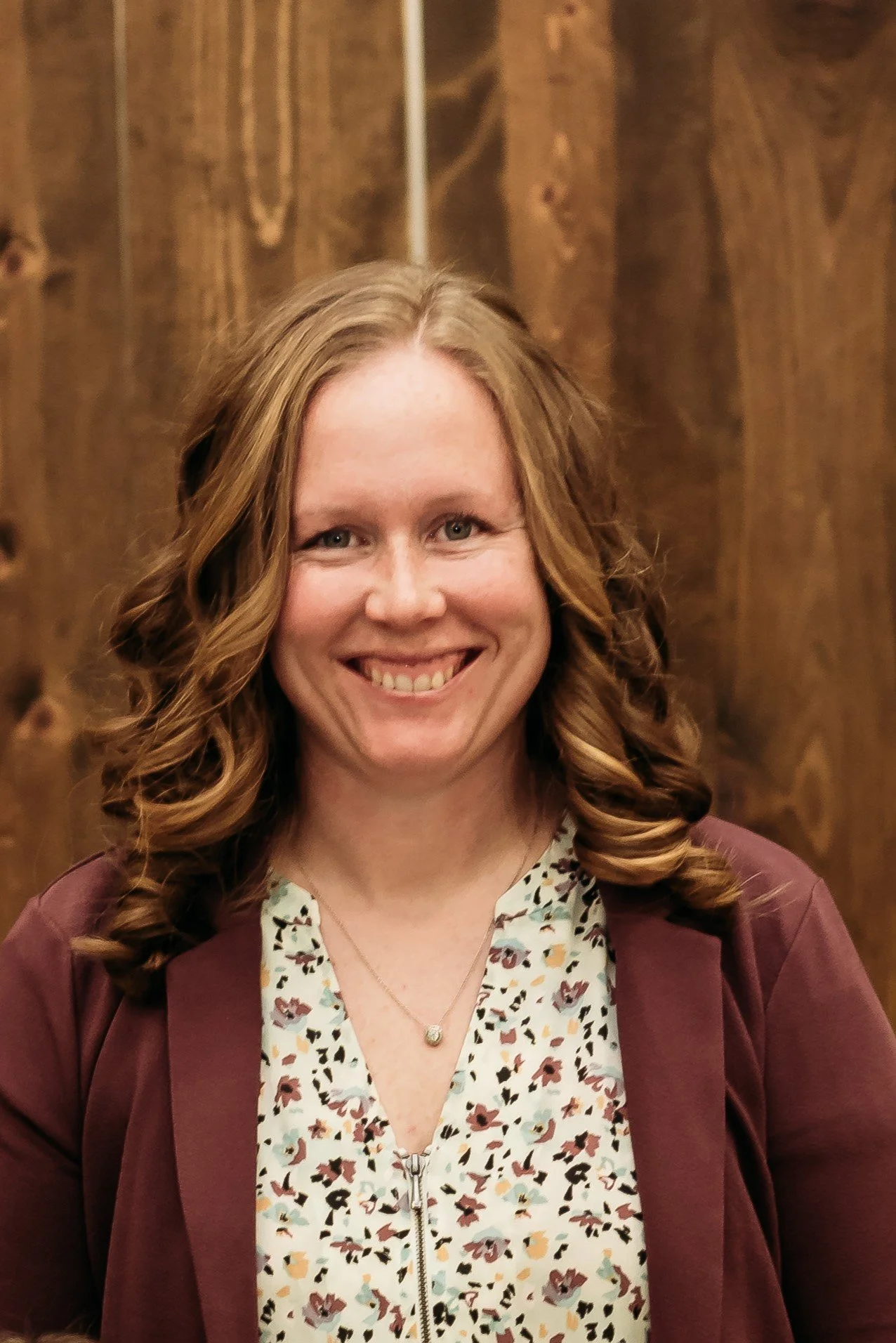 A woman with shoulder-length, curled red hair smiling, wearing a floral top and a burgundy blazer, standing in front of a wooden background.