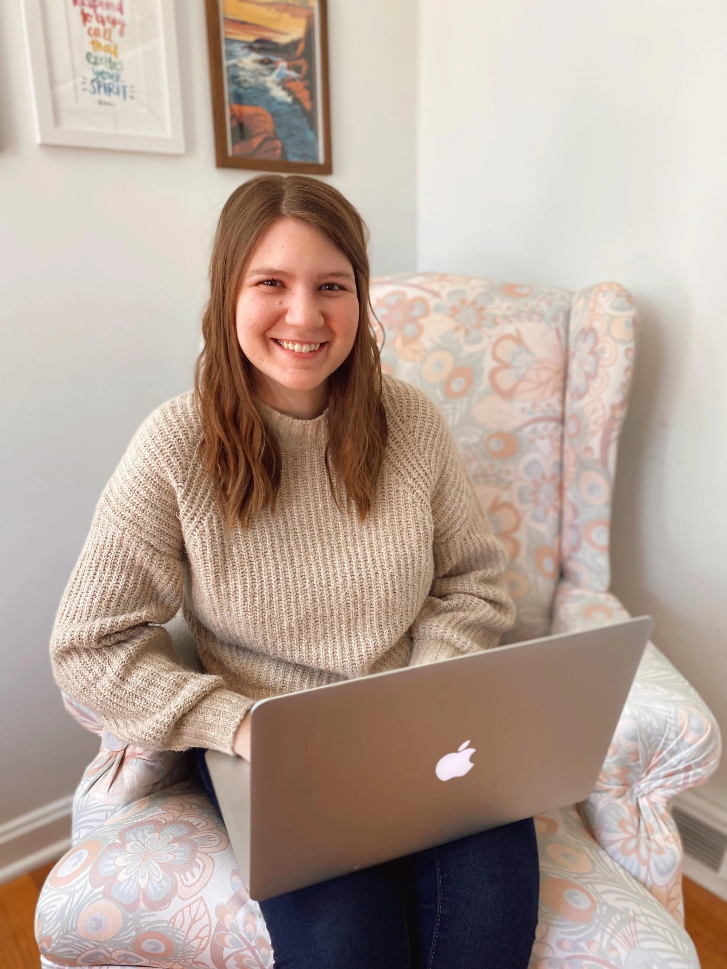 A young woman with shoulder-length brown hair smiling while sitting on a floral armchair with a laptop on her lap, in a room with framed artwork on the wall.