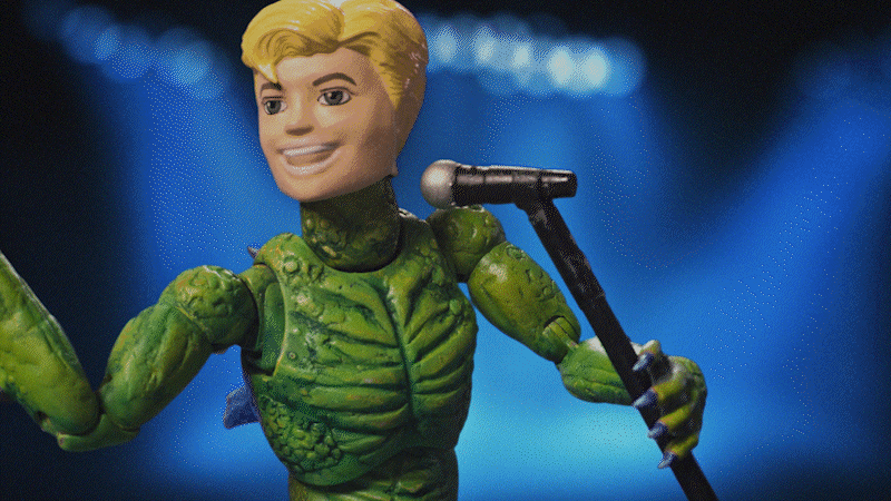 Animated toy figure with a human head and a green, insect-like body standing on stage with a microphone.