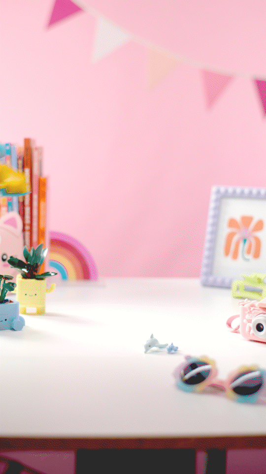 A decorated pink room with a white table, Stop motion animation of colorful toys, a frame with an orange flower design, and pink or pastel bunting hanging from the ceiling.