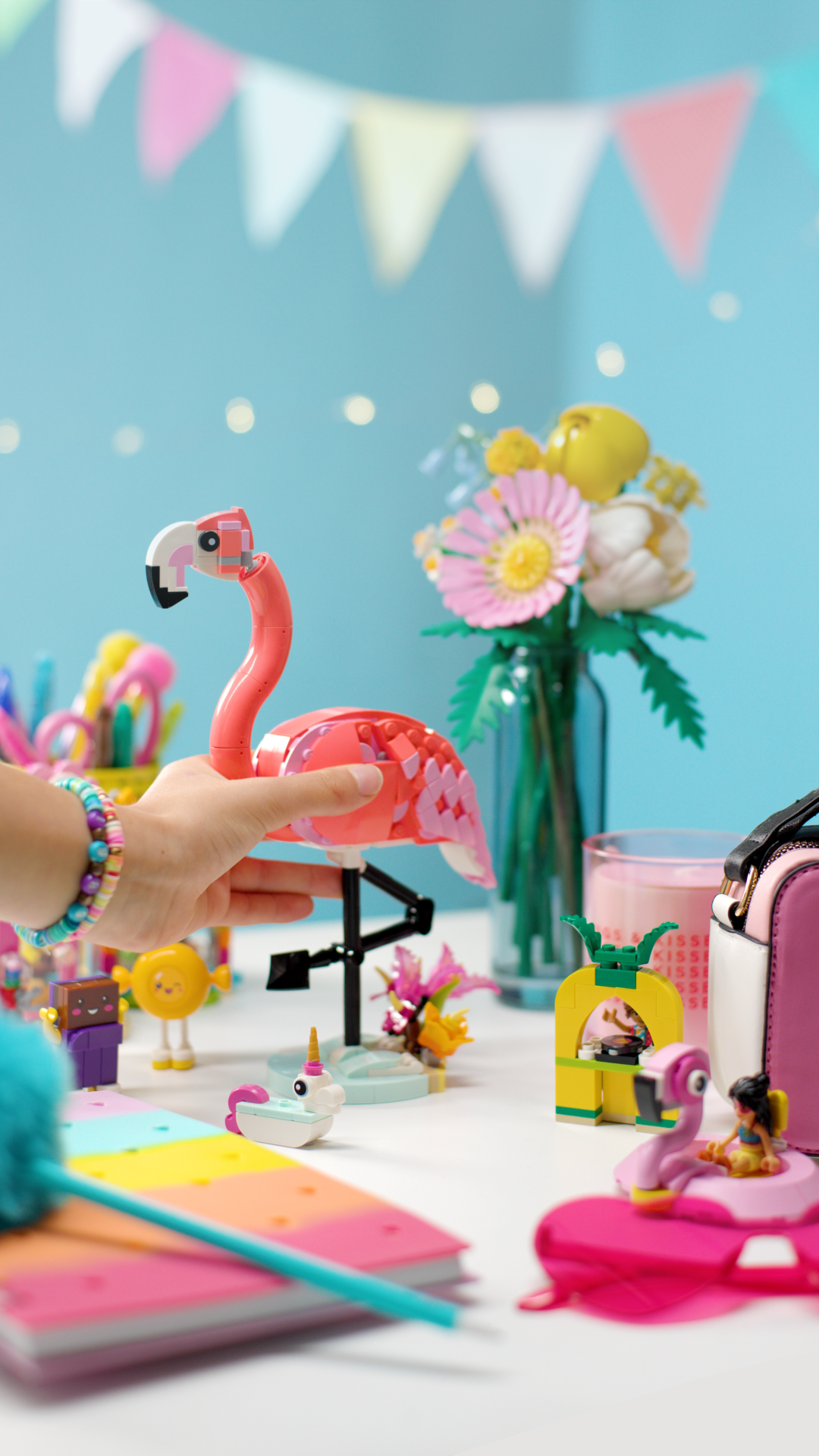 Colorful collection of toys and decorations on a white table, including a pink flamingo-shaped LEGO figure, a small yellow LEGO creature, a pink bag, a glass with a pink drink, colorful bunting, and a vase with flowers against a light blue background.