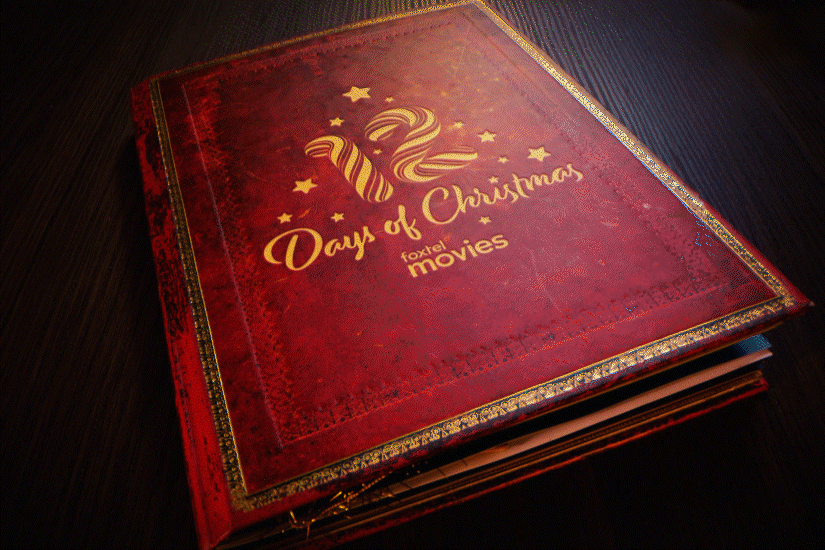 Stop motion animation of a red hardcover book titled '12 Days of Christmas', decorated with gold stars and a swirly number 12 on the cover, resting on a dark surface.