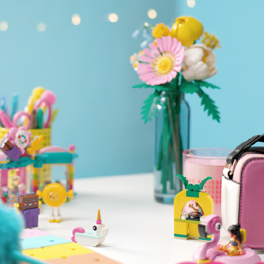 Stop motion animation of colorful toys, a pink flamingo, and a doll on a white surface with a blue wall and a vase of flowers in the background.