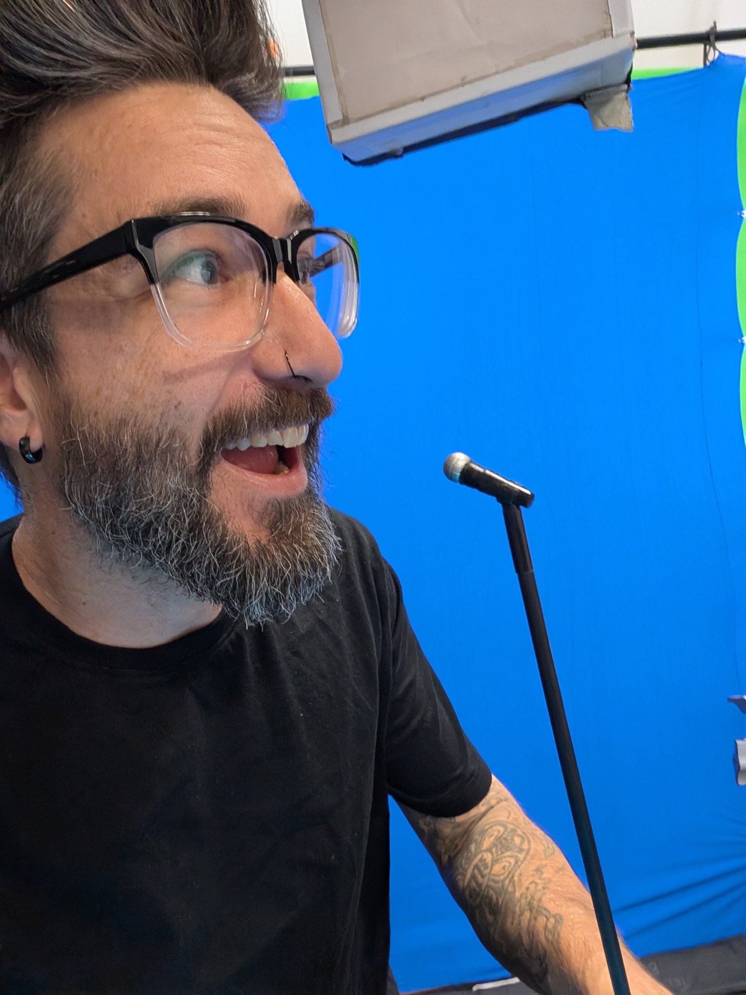 A man with glasses and a beard, smiling and speaking into a microphone with a blue background.