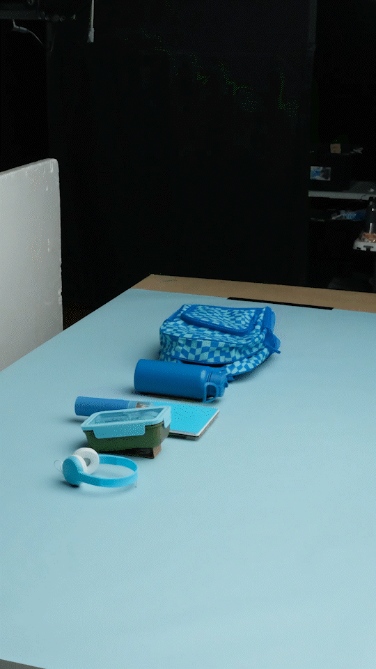 School supplies on a white table including a blue pencil case, a blue water bottle, a small notebook, a container with a pair of white earbuds, and a blue and white wallet.