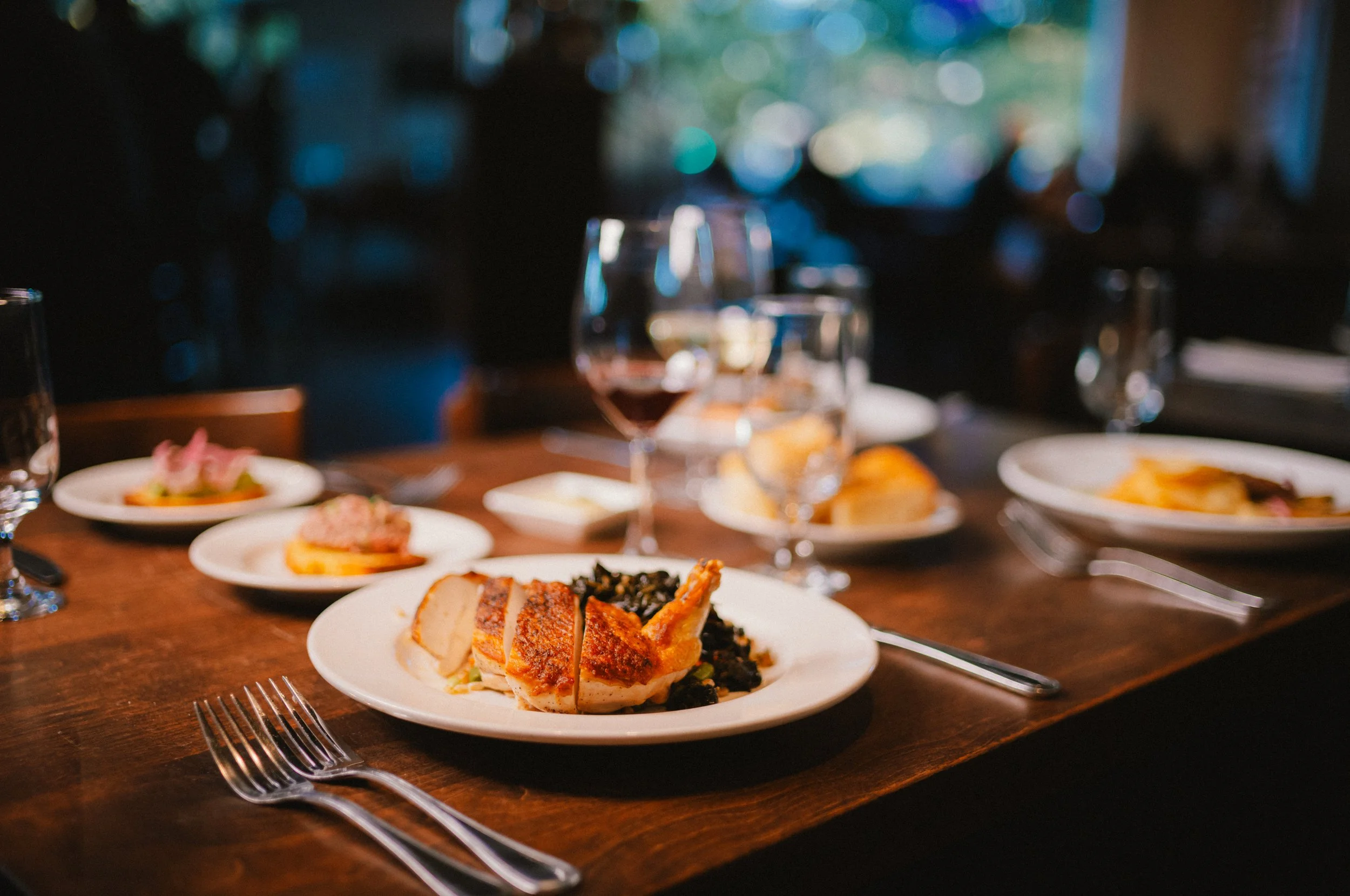 A dinner table set with various dishes, including a plate with sliced roasted chicken, and glasses of wine and water, in a dimly lit restaurant.