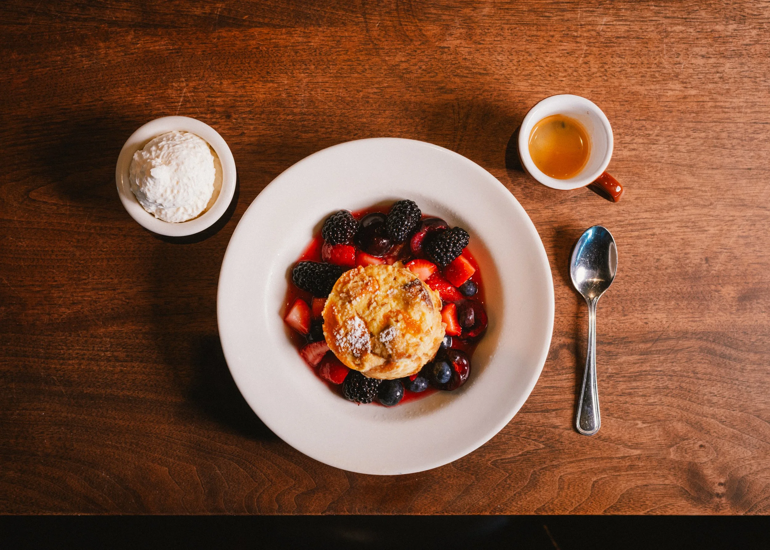 A plate with mixed berries and a baked apple topped with powdered sugar, served on a wooden table, accompanied by a spoon, a small cup of honey or syrup, and a small bowl of whipped cream.