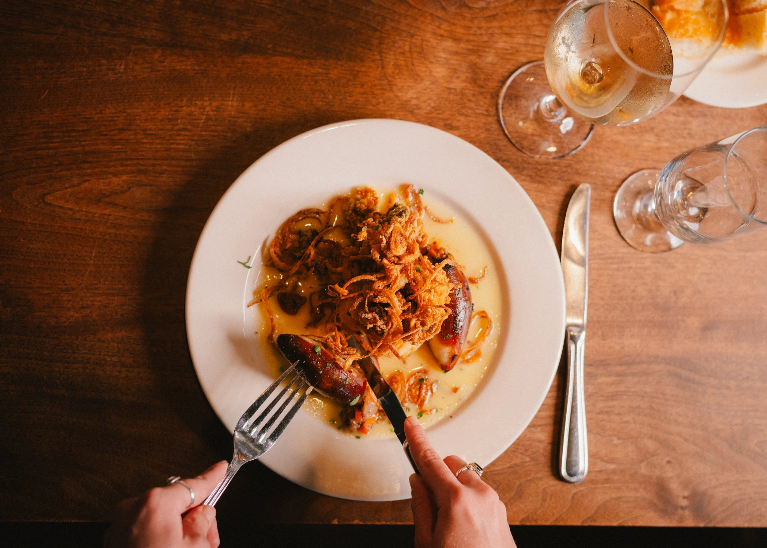 A person serving herself seafood, including lobster and crawfish, on a white plate at a wooden table with two glasses of champagne and a knife and fork.