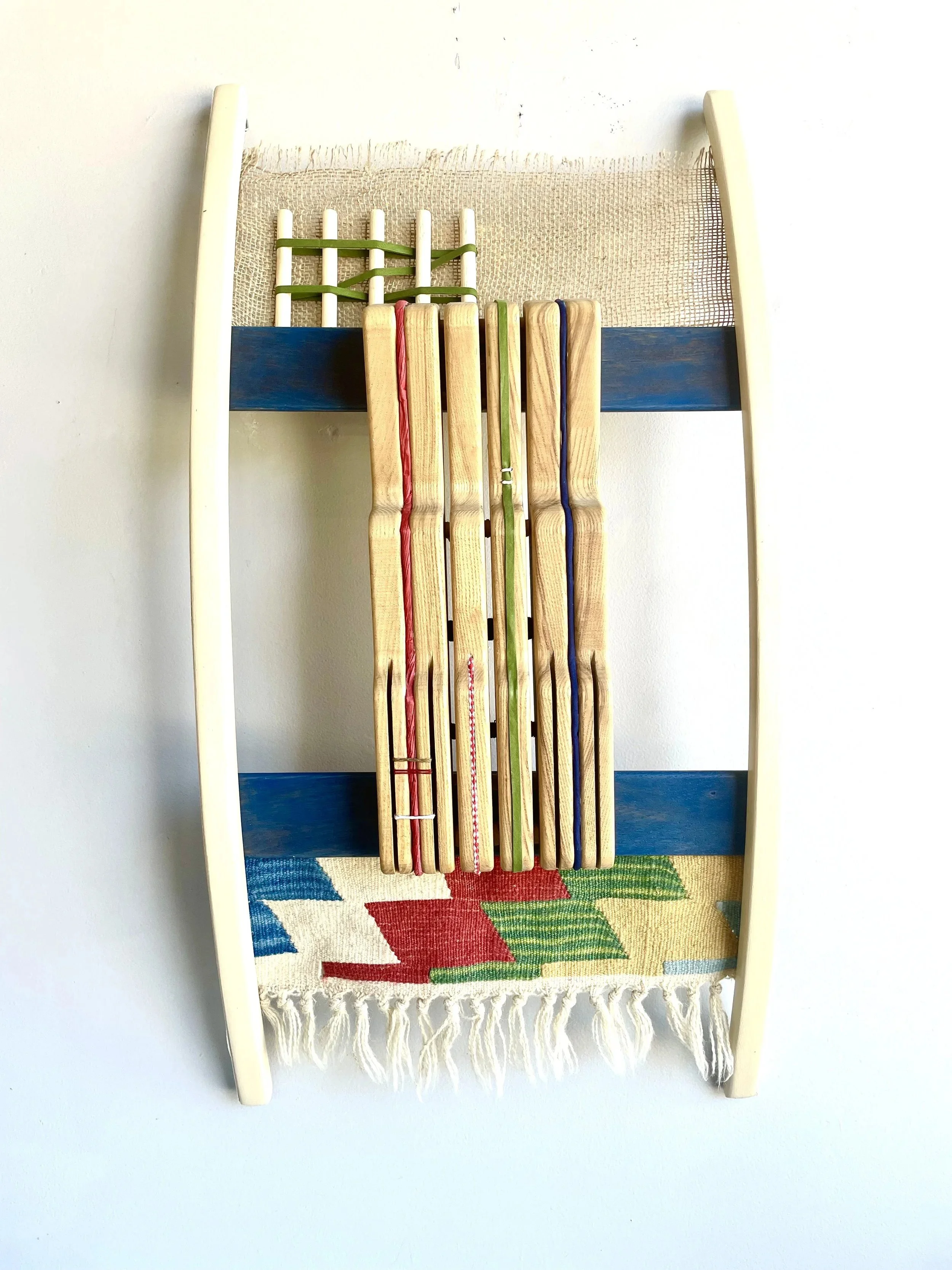 A wooden wall-mounted rack with blue horizontal supports, holding several wooden slats with colored strings, above a woven rug with a geometric pattern in red, green, blue, and white.
