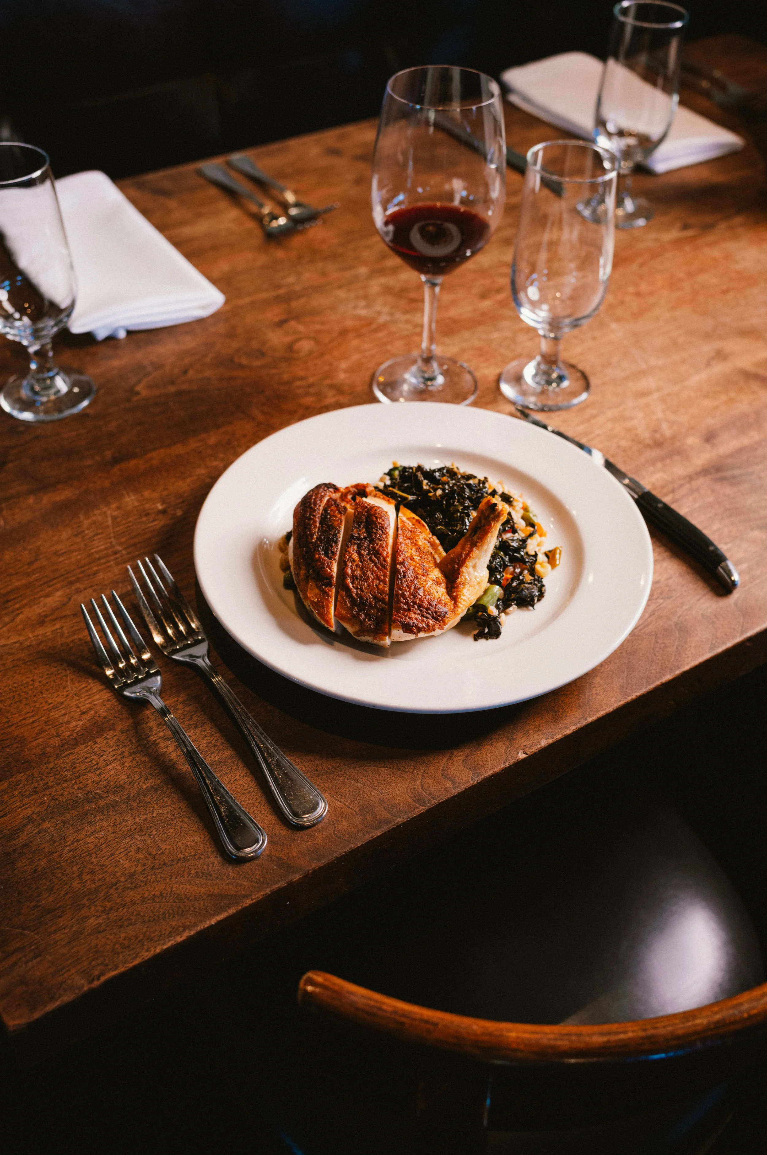 A dining table set with a white plate containing a sliced roasted chicken leg served with a bed of cooked greens, surrounded by glasses of red wine and empty water glasses, with silverware and folded white napkins.