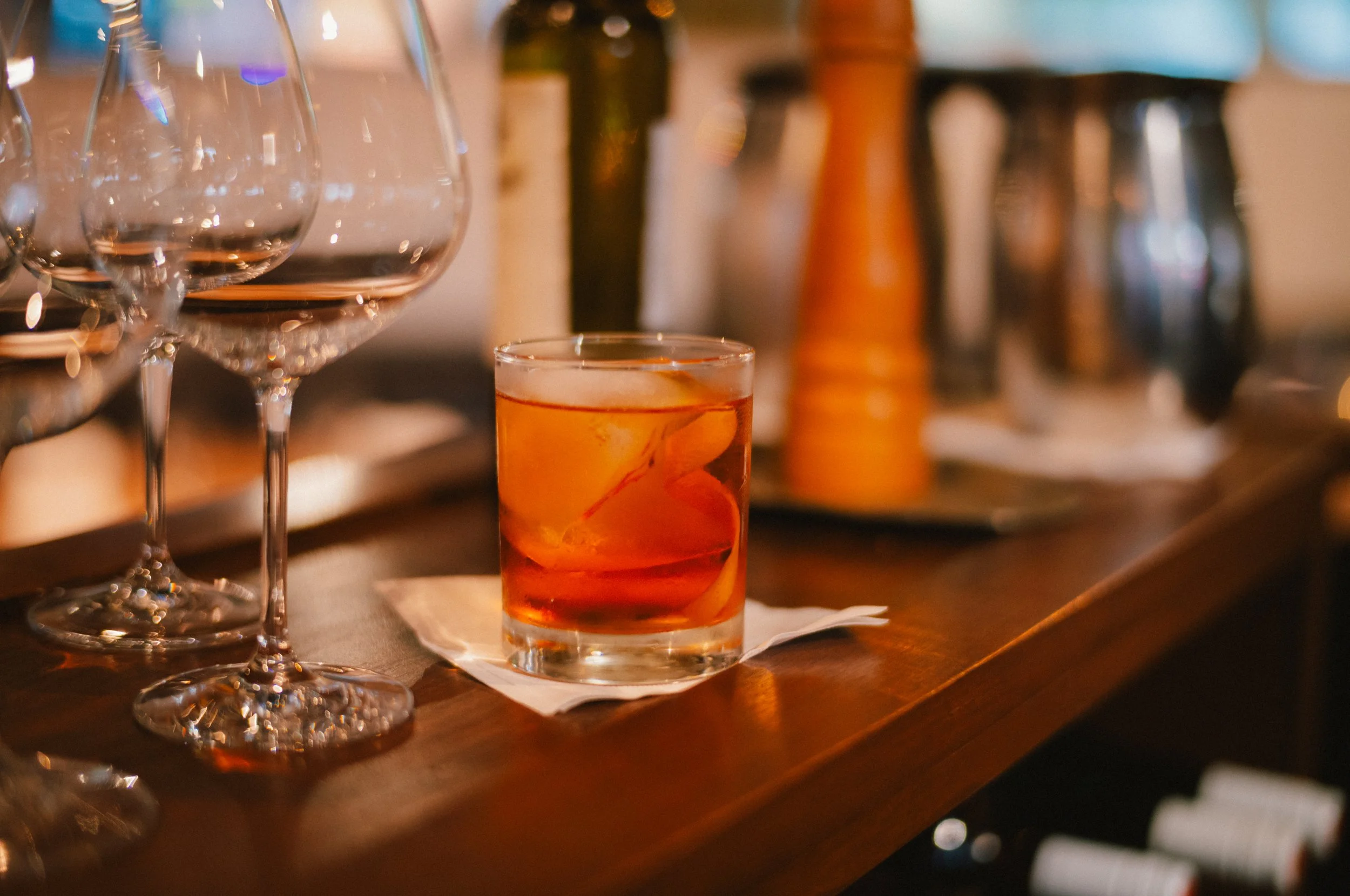 A glass of amber-colored drink with ice and orange slices on a white napkin on a wooden bar counter, with several wine glasses and bottles blurred in the background.
