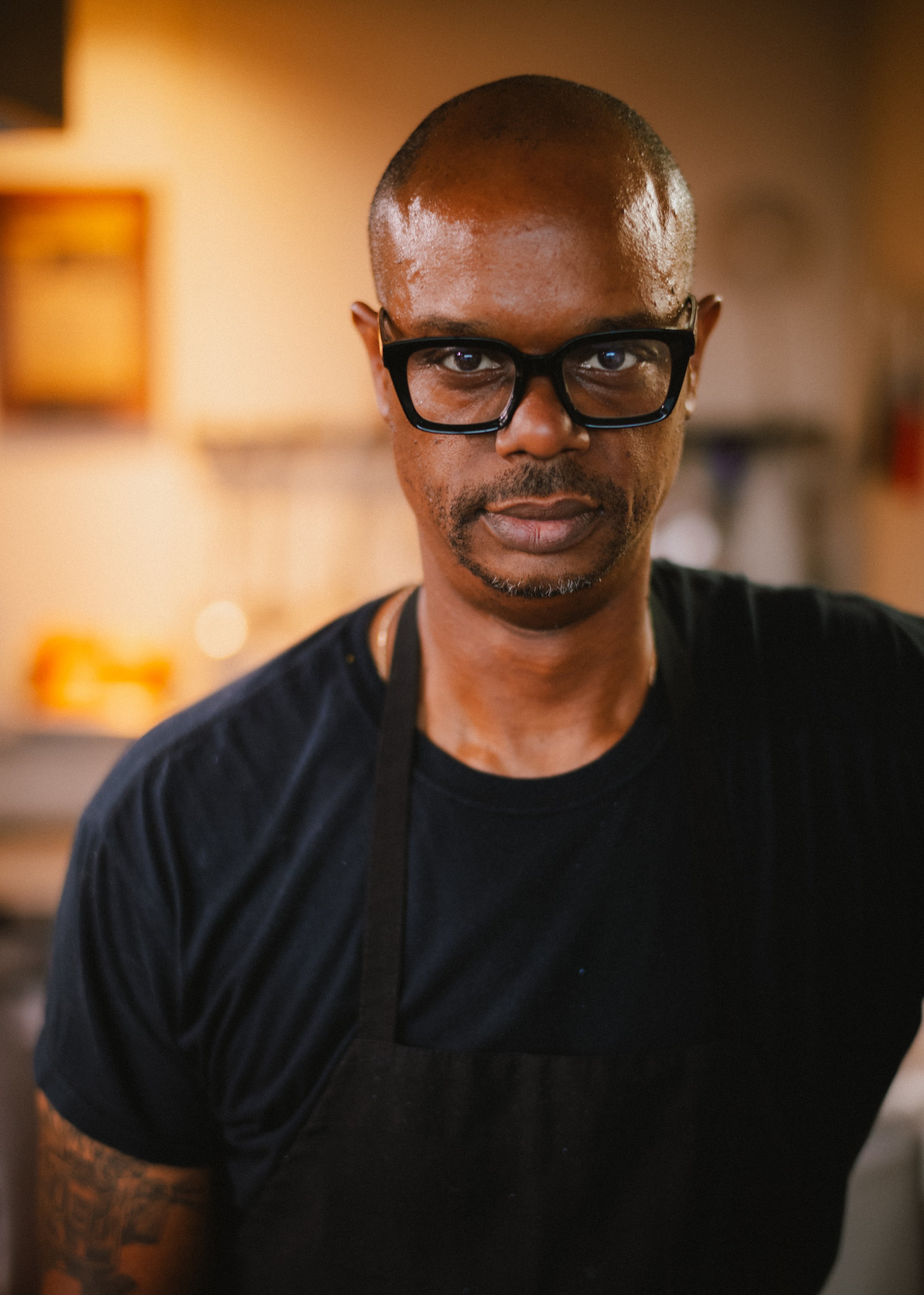 A man with short hair, wearing glasses and a black T-shirt, standing in a warmly lit room with blurred background.