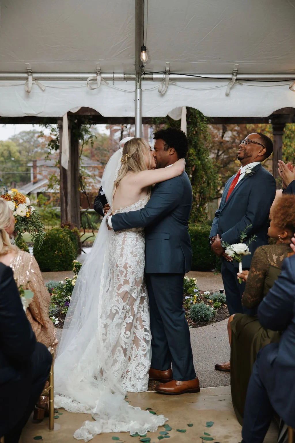 A bride and groom kiss during their outdoor wedding ceremony under a white tent, surrounded by seated guests holding flowers, with trees in the background.