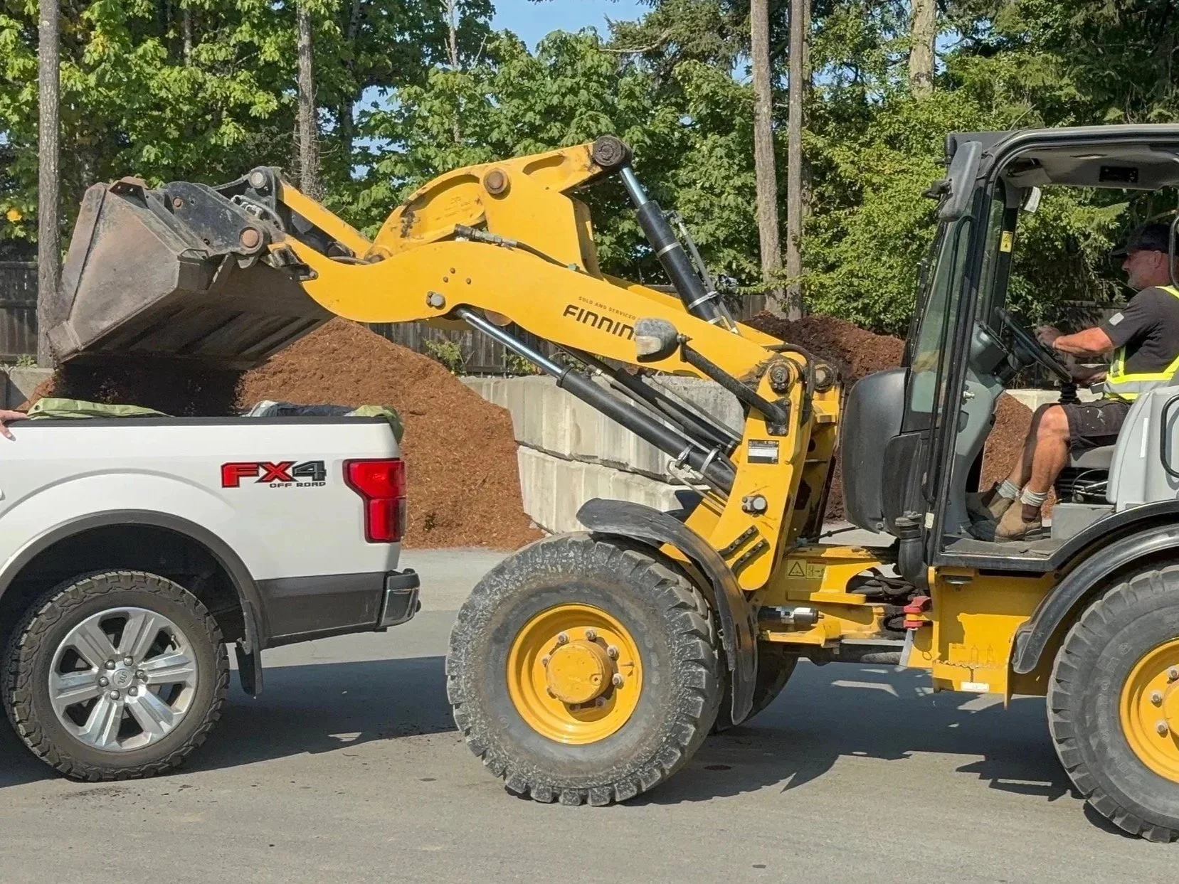 Construction vehicle loading dirt into a pickup truck on a road with a wooded background.