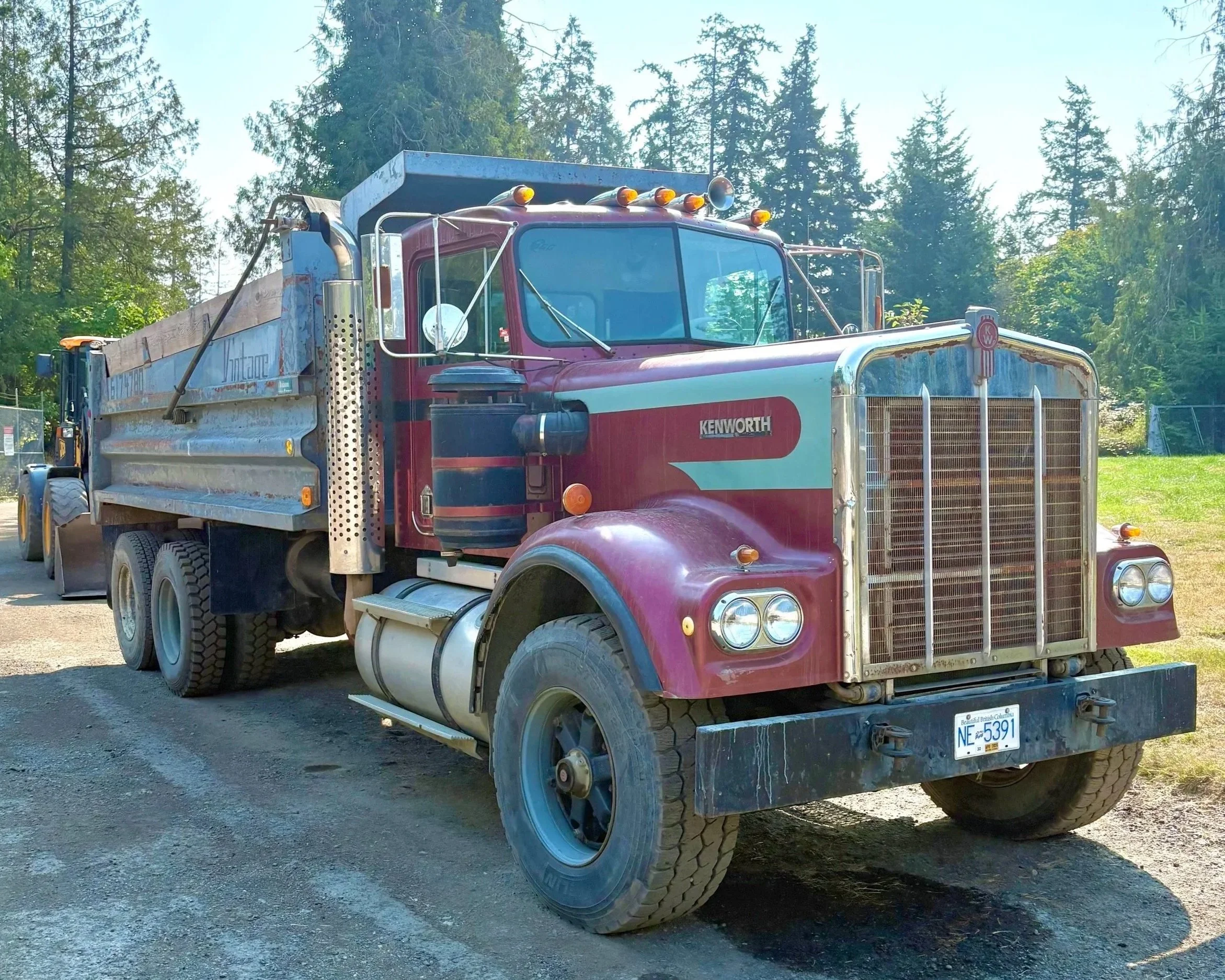 A red Kenworth dump truck parked on a gravel surface with a wooded background.