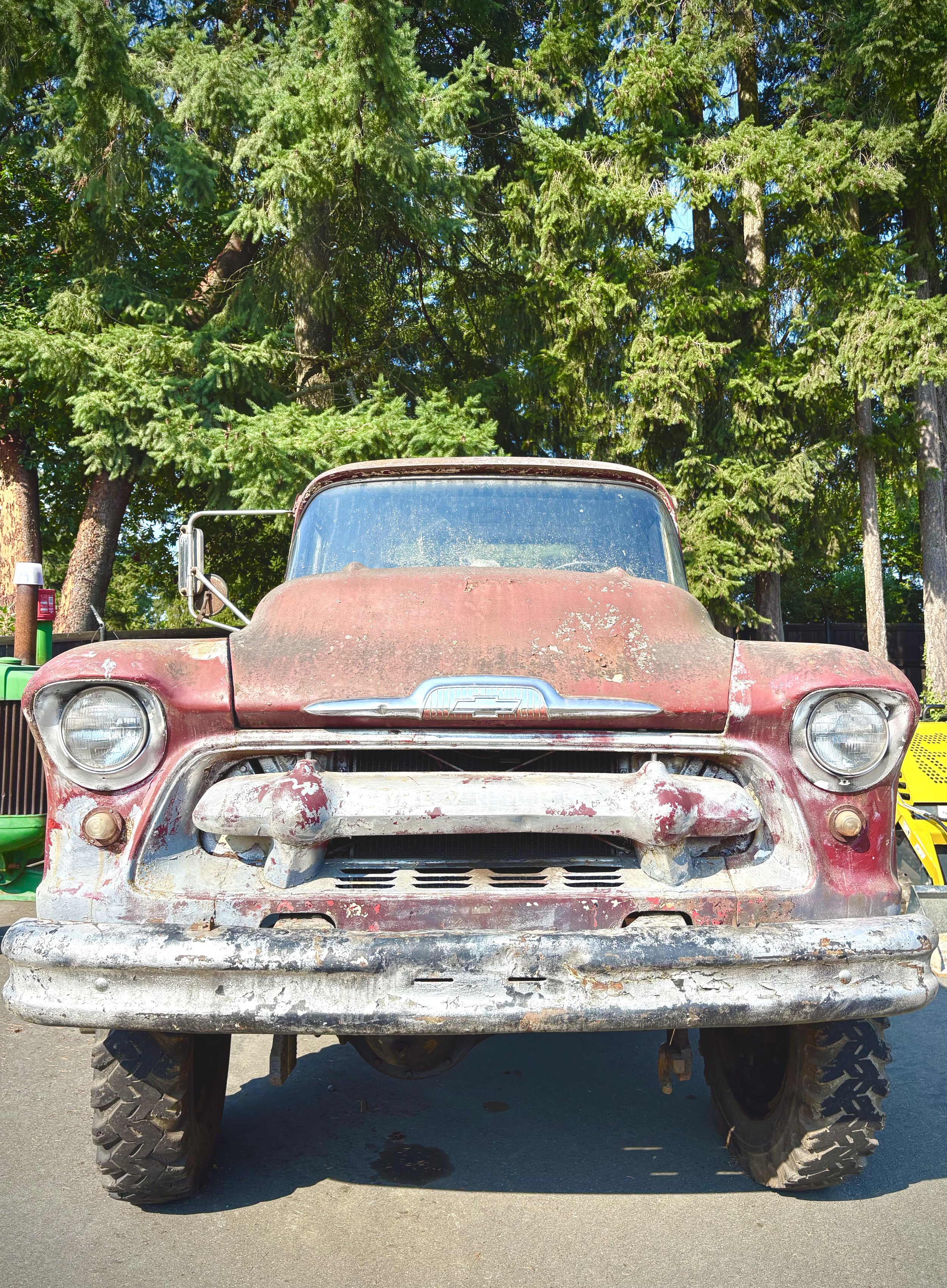 An old, rusty red pickup truck with a weathered front bumper, parked outdoors in a wooded area with tall green trees in the background.