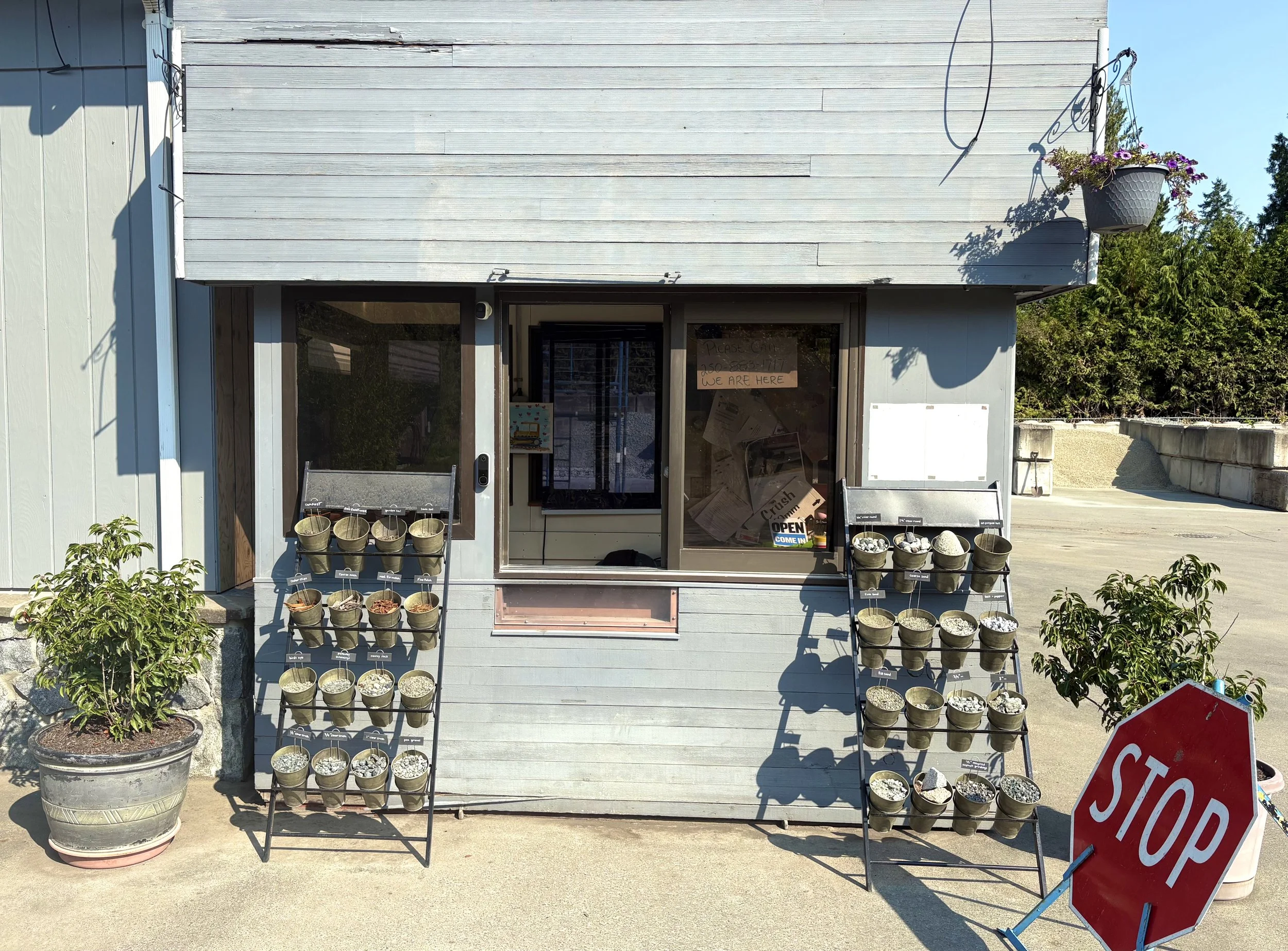 A small storefront with two display racks of potted plants outside. The store has a sign on the window and a stop sign in front.