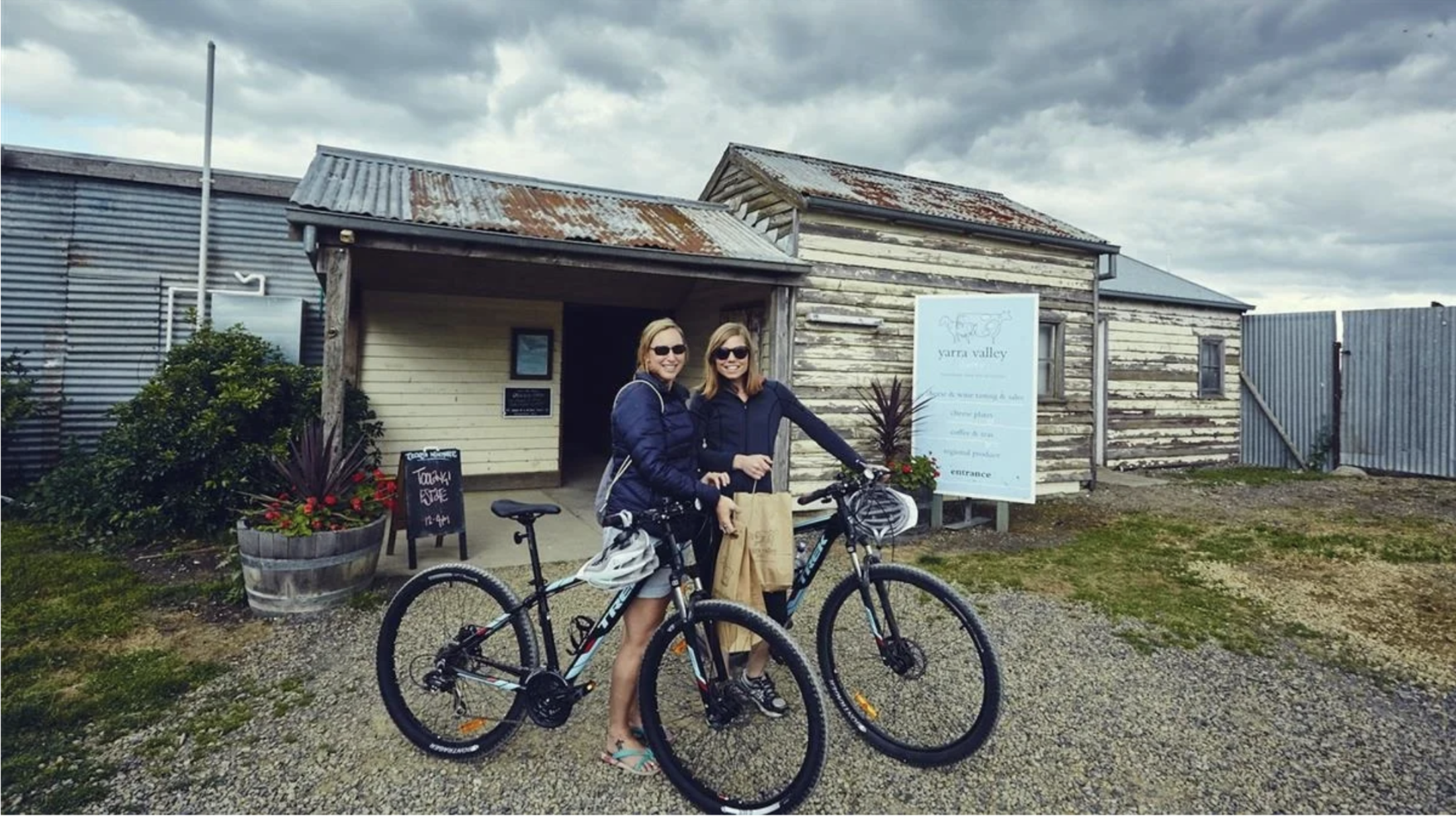 Two women with sunglasses smiling, standing with bicycles in front of a rustic wooden building with a sign and potted plants, on a cloudy day.