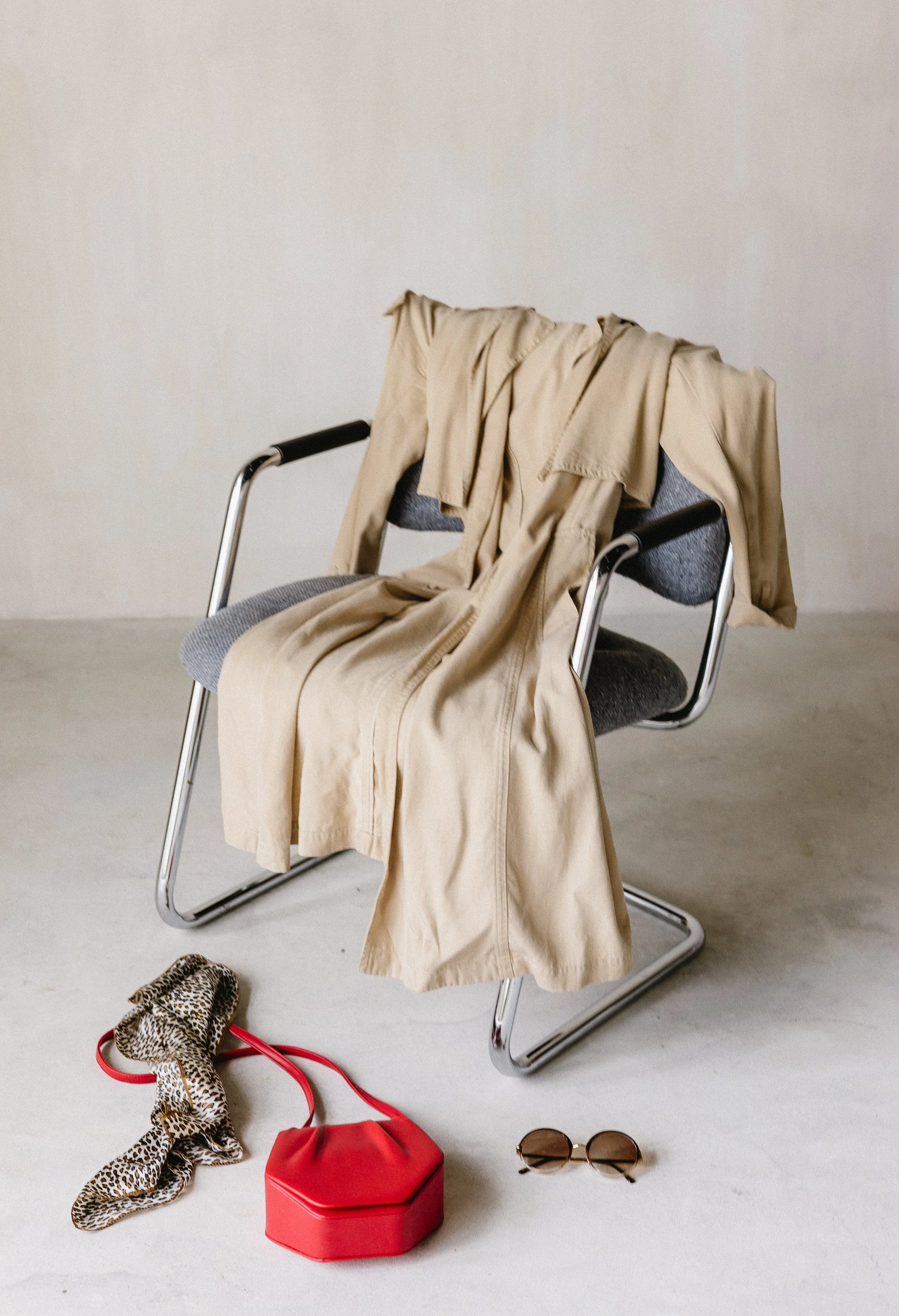 A modern office chair with a beige coat and a light brown scarf draped over it, placed in a minimalistic room. On the floor in front, there's a leopard print scarf, red purse, and round sunglasses.