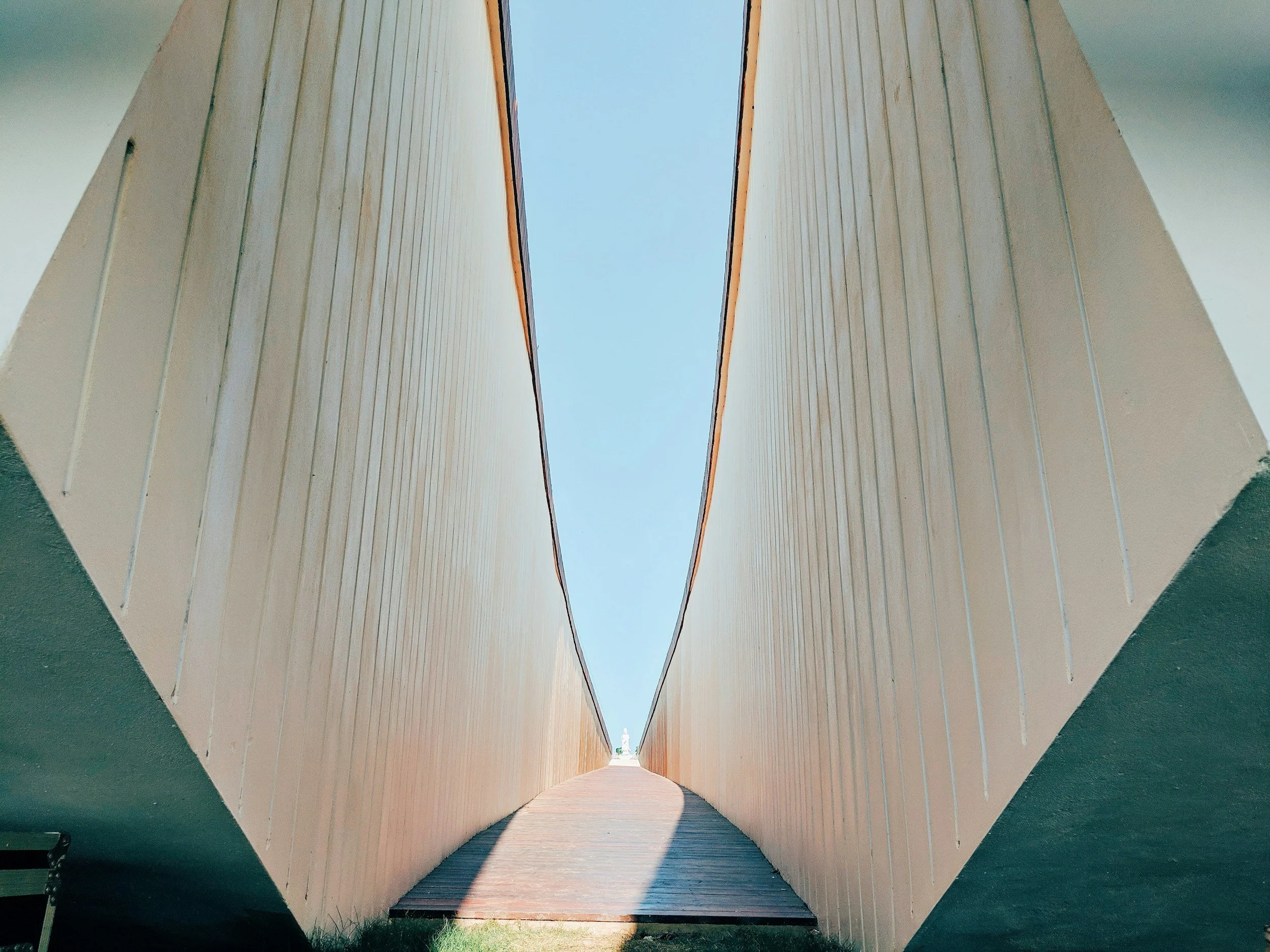 A modern architectural structure with two large beige walls forming a narrow V shape, opening to a clear blue sky, with a wooden pathway leading into the distance.