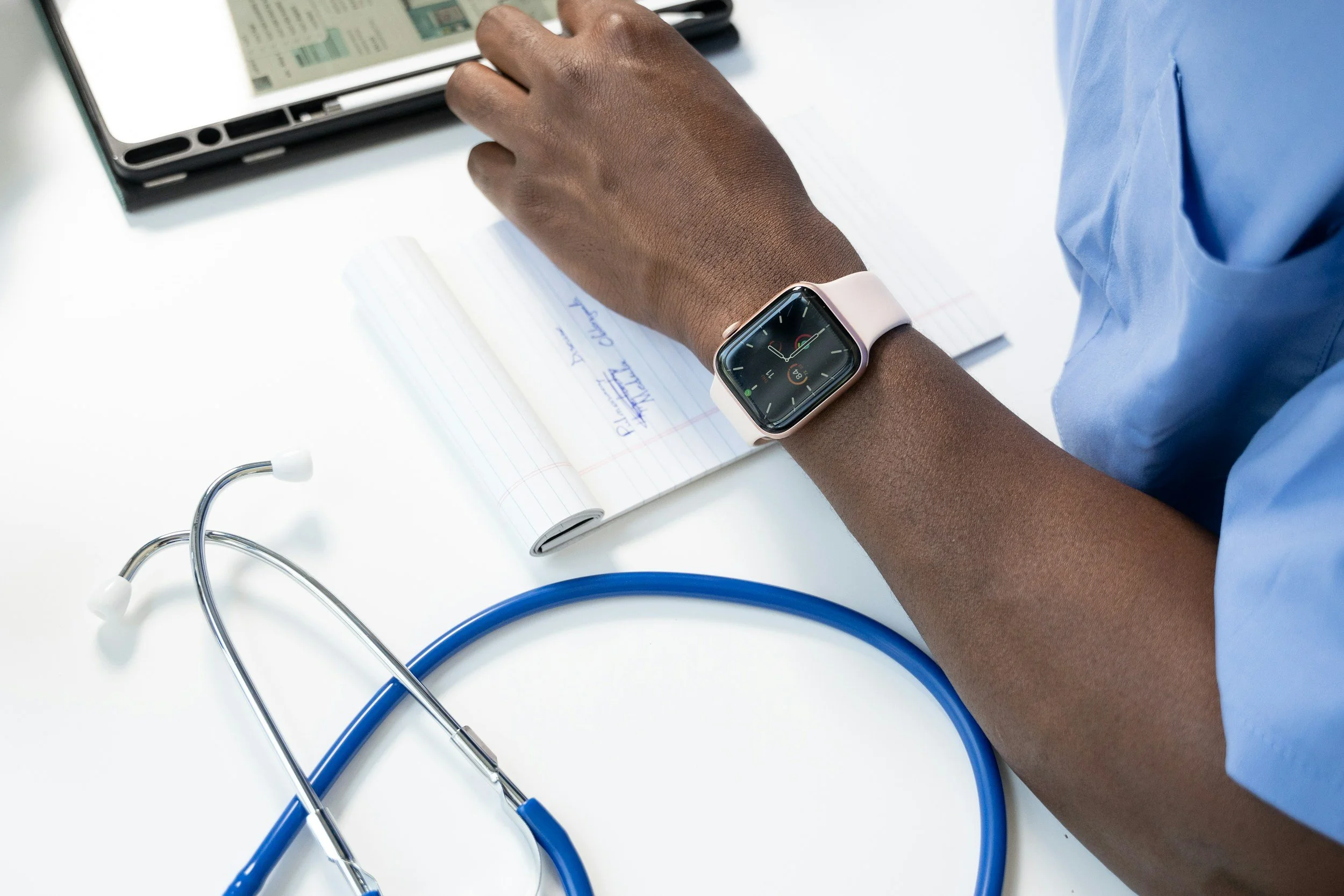 A healthcare professional wearing a smartwatch sitting at a white desk with a stethoscope, a tablet, and a notepad.