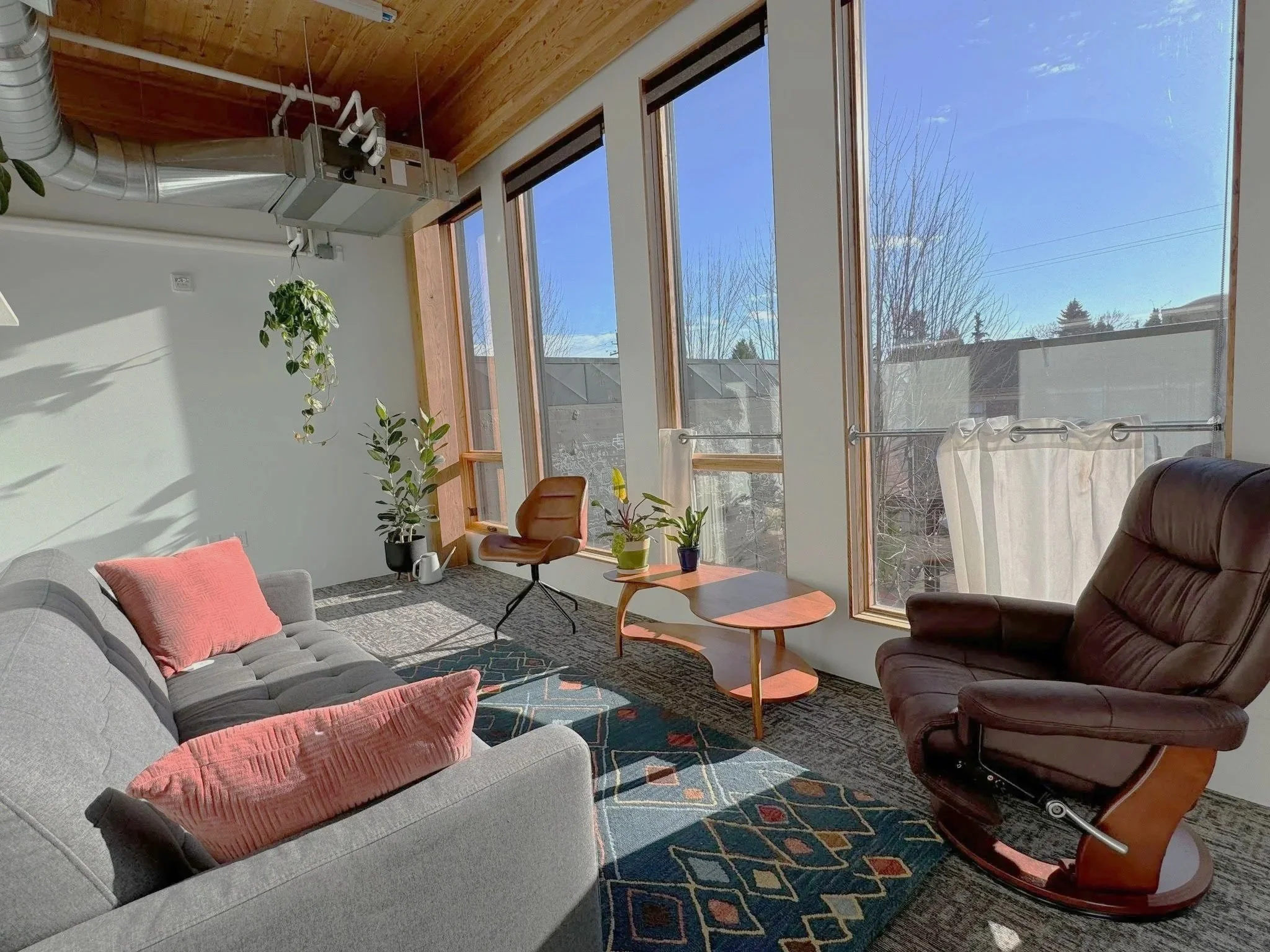 A bright therapy room for retune space offering therapy in Portland, OR  with large windows, a gray sofa with pink cushions, a brown leather armchair, a wooden coffee table with plants, and potted plants near the windows, with sunlight streaming in.