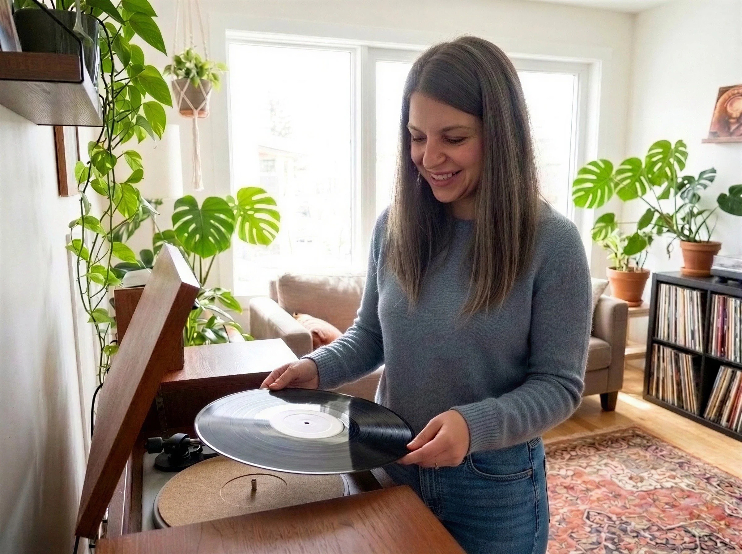 Young woman with brown hair smiling while handling a vinyl record on a turntable in a bright living room with potted plants and a bookshelf.
