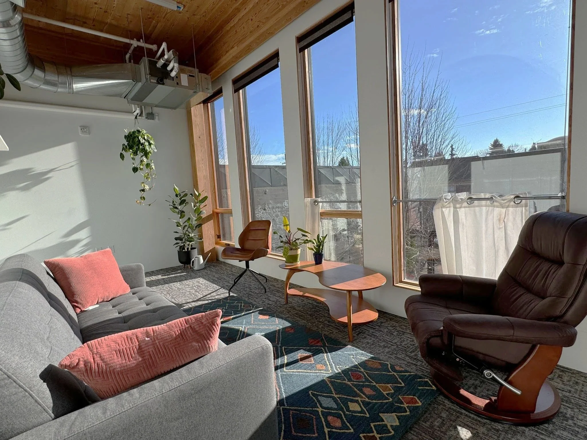 A modern living room with large windows, a gray sofa with pink pillows, a brown leather armchair, a wooden coffee table, potted plants, and natural sunlight.