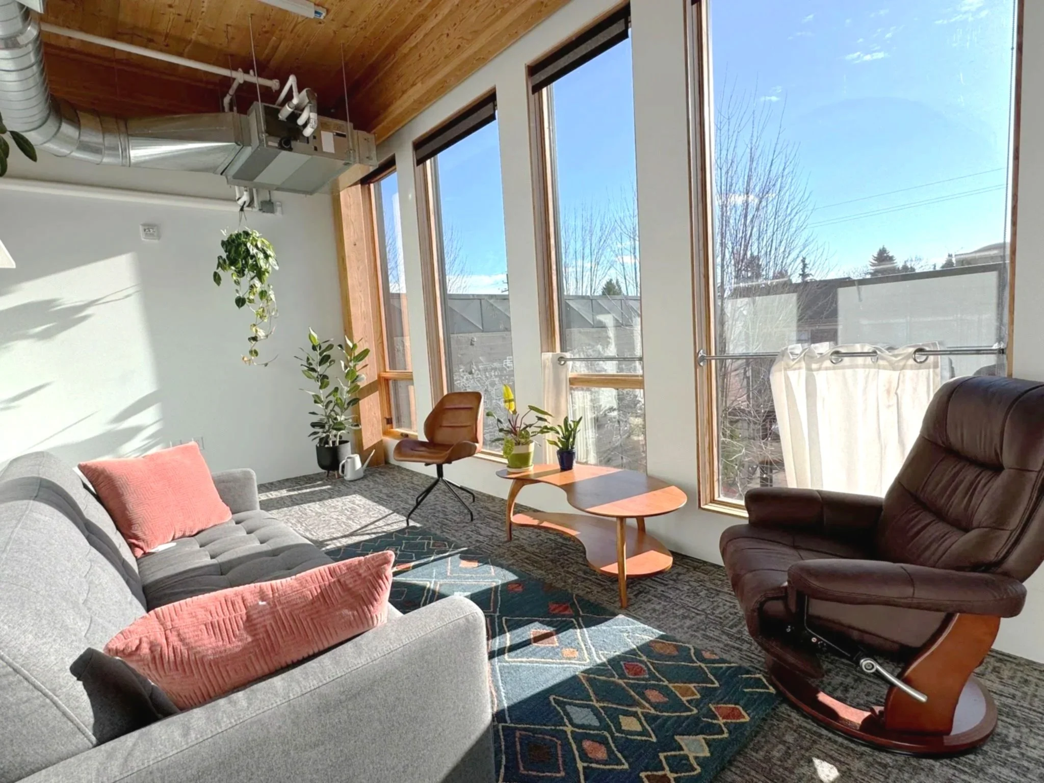 Bright living room with large windows, gray sofa with pink cushions, wooden chair, small coffee table with plants, and a brown leather recliner, with sunlight streaming in.
