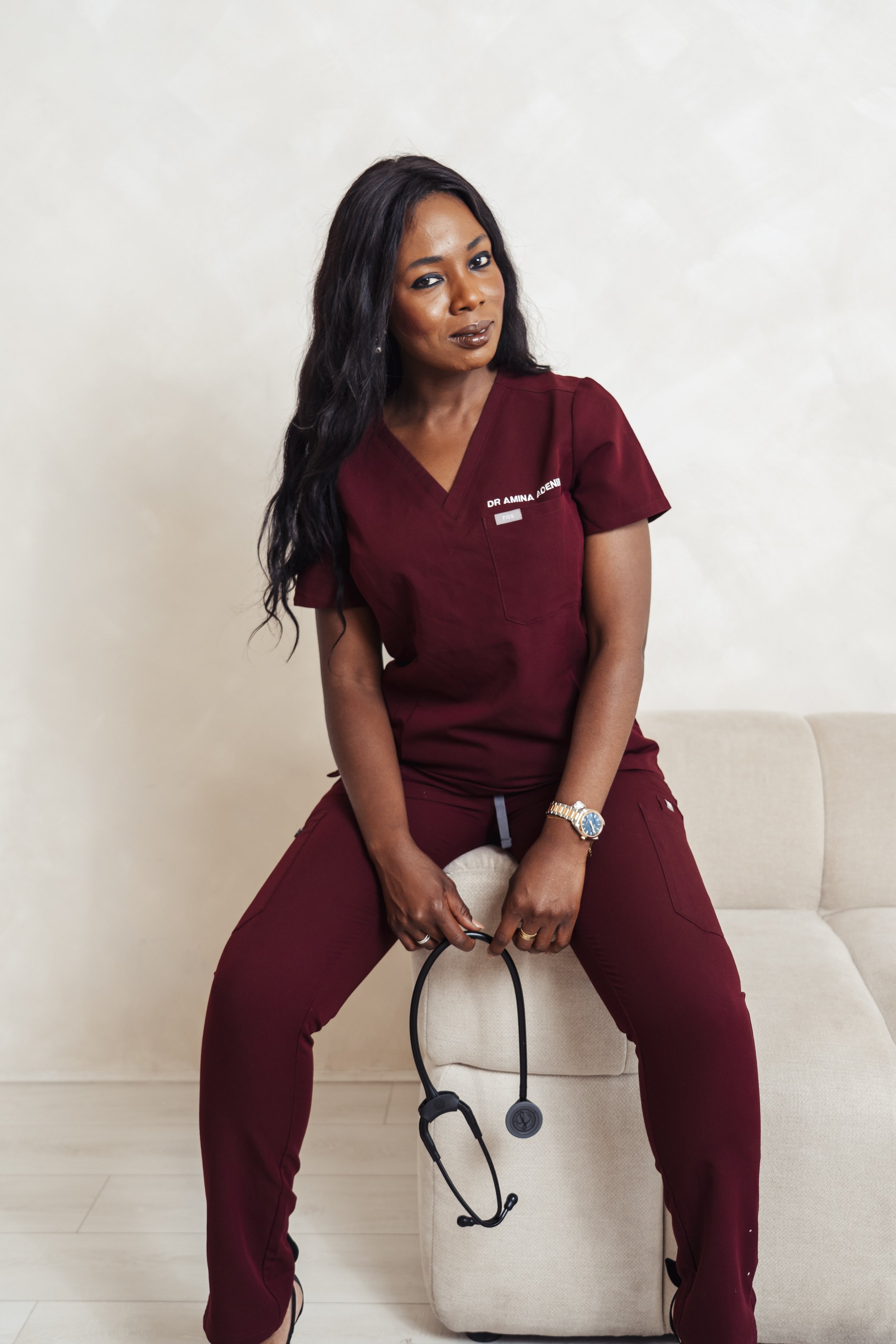 African American woman in burgundy medical scrubs, sitting on a small beige ottoman, holding a stethoscope, with a confident expression, in a medical office setting.
