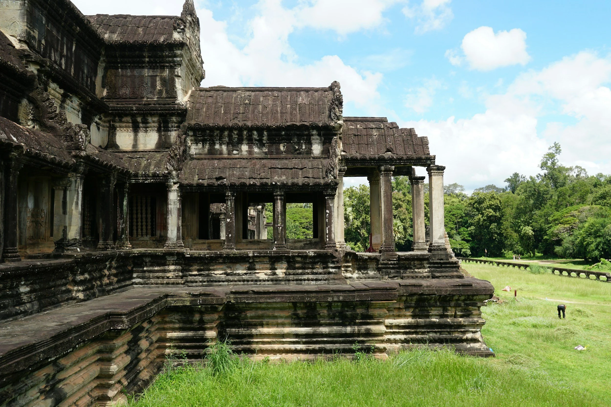 Ancient stone temple ruins with multiple levels, columns, and a tiled roof, surrounded by green grass and trees under a partly cloudy sky.