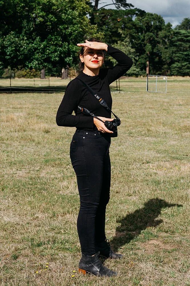 On location portrait of Dijana. Candid photo of Dijana holding camera and looking back towards camera. She wears all black, red lipsick and is smiling warmly. Her hand covers her forehead to block the bright sun. She is standing in a green feild.