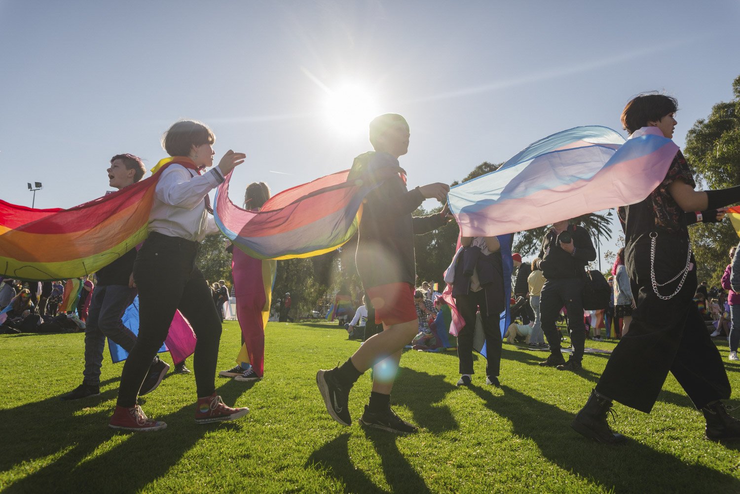Midsumma Pride March Photography of a group of people gather on an oval getting ready to go to Pride March, St Kilda. 3 queer youth parade in a line, wearing rainbow and trans pride flags, the sun glowing behind them