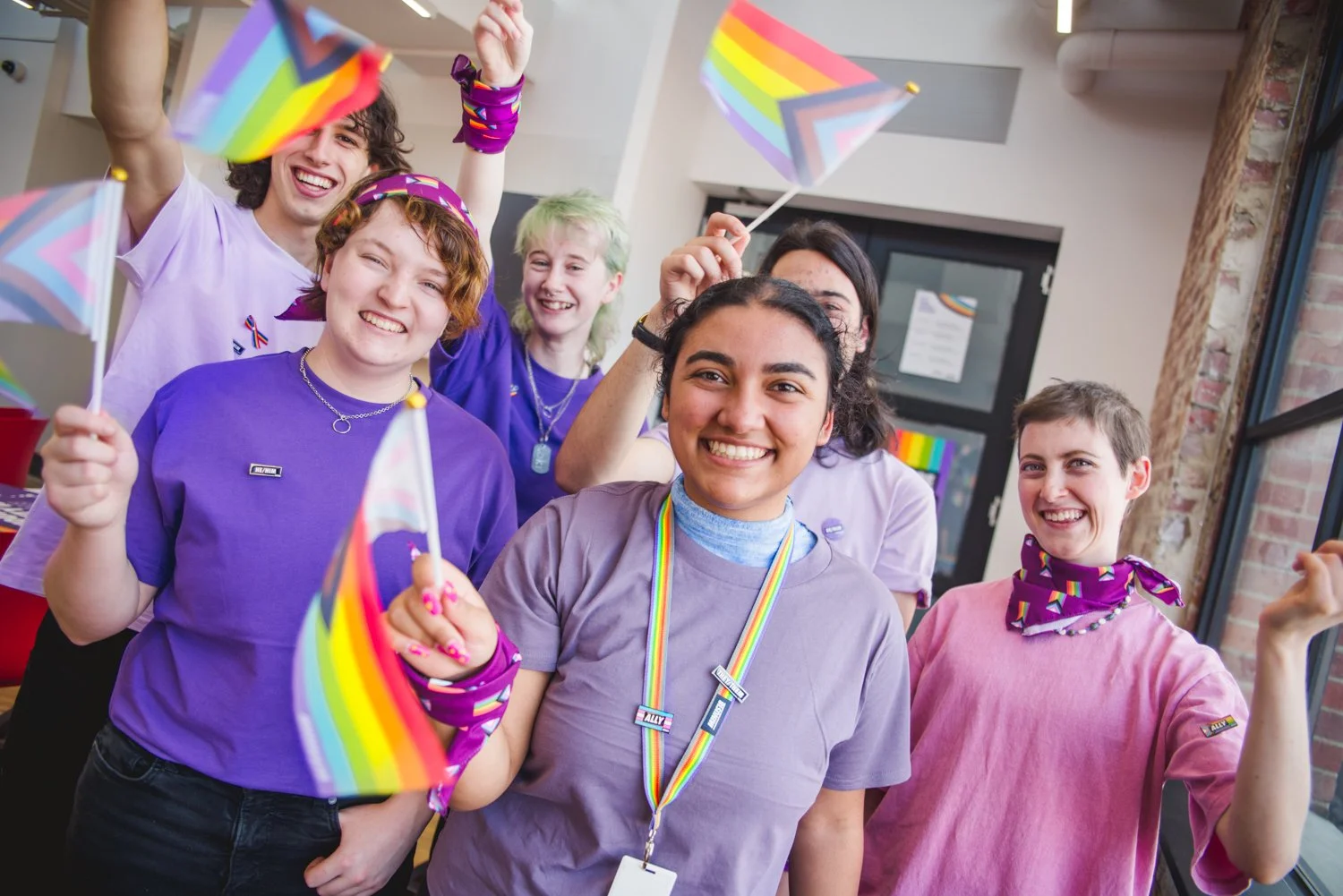 5 young people waving the rainbow flag and wearing purple to celebrate Wear it Purple day. Everyone looks happy.